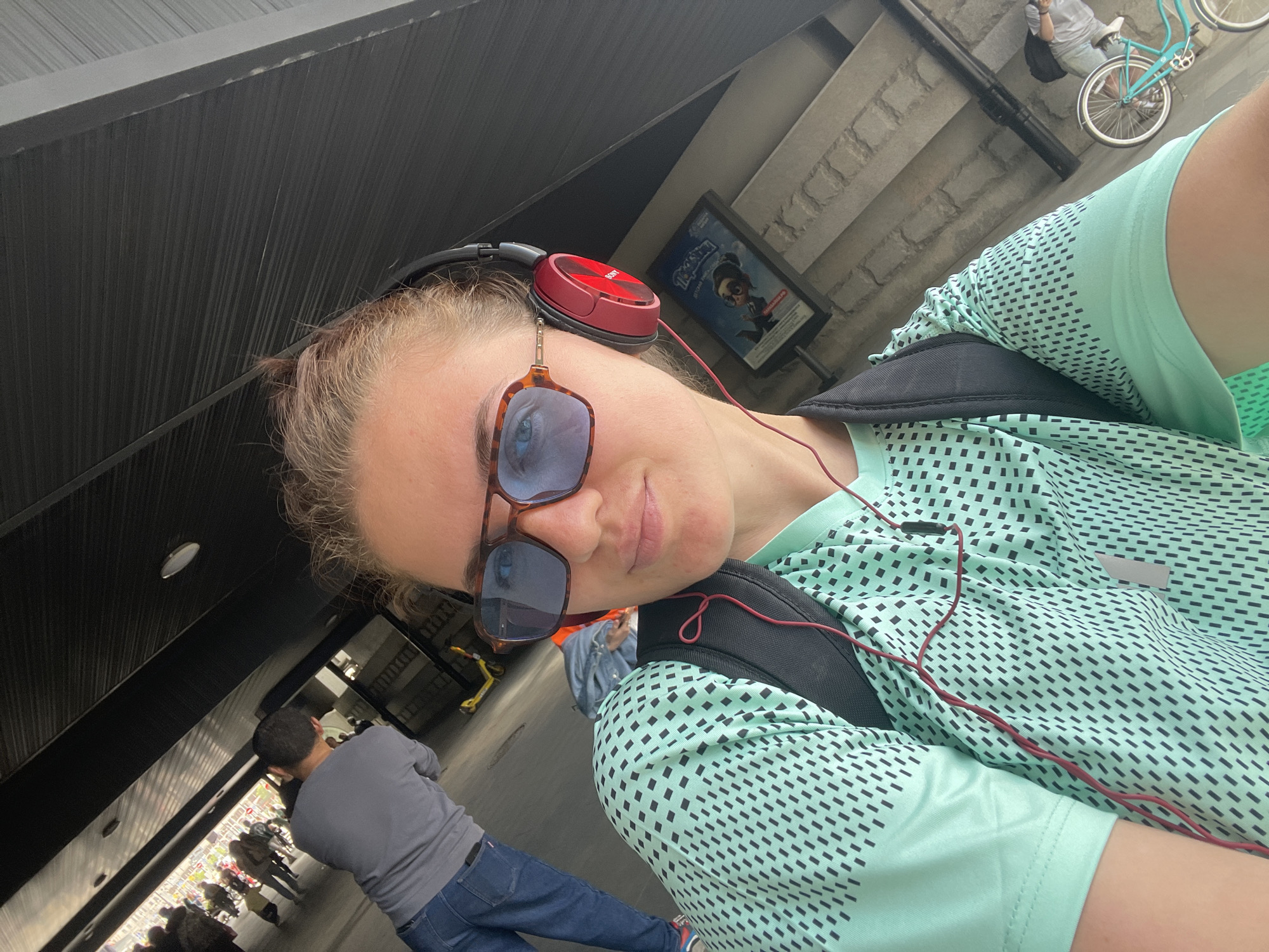 A person with fair skin is the primary subject of this eye-level selfie. The person is wearing red headphones and tinted sunglasses. They have a black backpack and a light green, patterned shirt. The photo shows a portion of the person's face, shoulder, and chest.
In the background, other people are walking on a pedestrian walkway. A person in a gray shirt and blue jeans is walking in the same direction as the selfie-taker. Above and to the side of the selfie-taker is a poster and an outdoor staircase with a bicycle parked on it.
Based on the poster's Cyrillic text and style, this is potentially in Moscow, Russia.