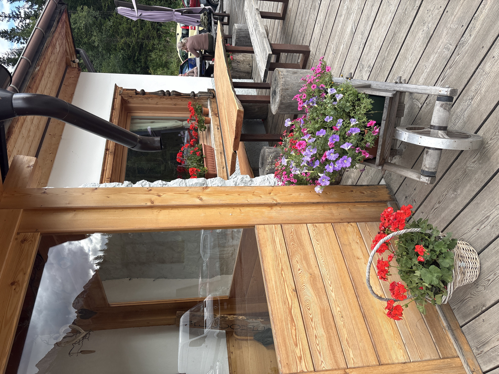 The image captures an outdoor seating area of a building, likely a cafe or restaurant, situated in Auronzo di Cadore, Italy. The foreground features a weathered wooden deck, upon which a rustic wicker basket overflows with vibrant red geraniums. To the right, a wooden garden cart holds a lush arrangement of purple and pink petunias.
Beyond the flowering plants, a large, rough-hewn wooden table is set up with matching benches and tree stump-like stools. Further back, a person is seated at a table, facing away from the camera. The building itself has a white facade and wooden trim around its windows. A dark brown downspout runs along the roofline.
Reflections in the large window of the building show a cloudy sky and distant pine trees, suggesting a mountainous or forested environment typical of the Italian Alps. In the background, parked vehicles are visible, including a white van, a blue car, and other cars. The overall scene conveys a relaxed, rustic atmosphere, characteristic of an alpine establishment.