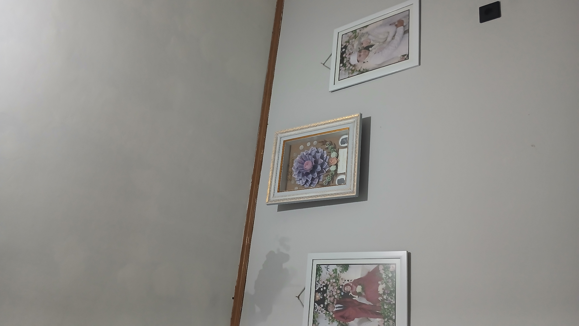 The image shows three framed pieces of art hanging on a light grey wall. The artwork consists of two framed photographs and one framed shadow box. The photograph on the left depicts a couple dressed in formal attire, presumably for a wedding, with the male in a maroon suit and the female in a maroon dress. The shadow box in the center contains a decorative arrangement resembling a flower, made from what appears to be currency, with small silhouette cutouts at the bottom. The photograph on the right also features a couple in formal wedding attire, both wearing white. Above the artwork, a wooden trim runs horizontally along the wall. The scene is illuminated by an unseen light source, casting subtle shadows on the wall. A small black electrical outlet cover is visible on the wall to the right of the central artwork. The location of this scene within Sampang, Indonesia, is not determinable from the visual information provided in the image.