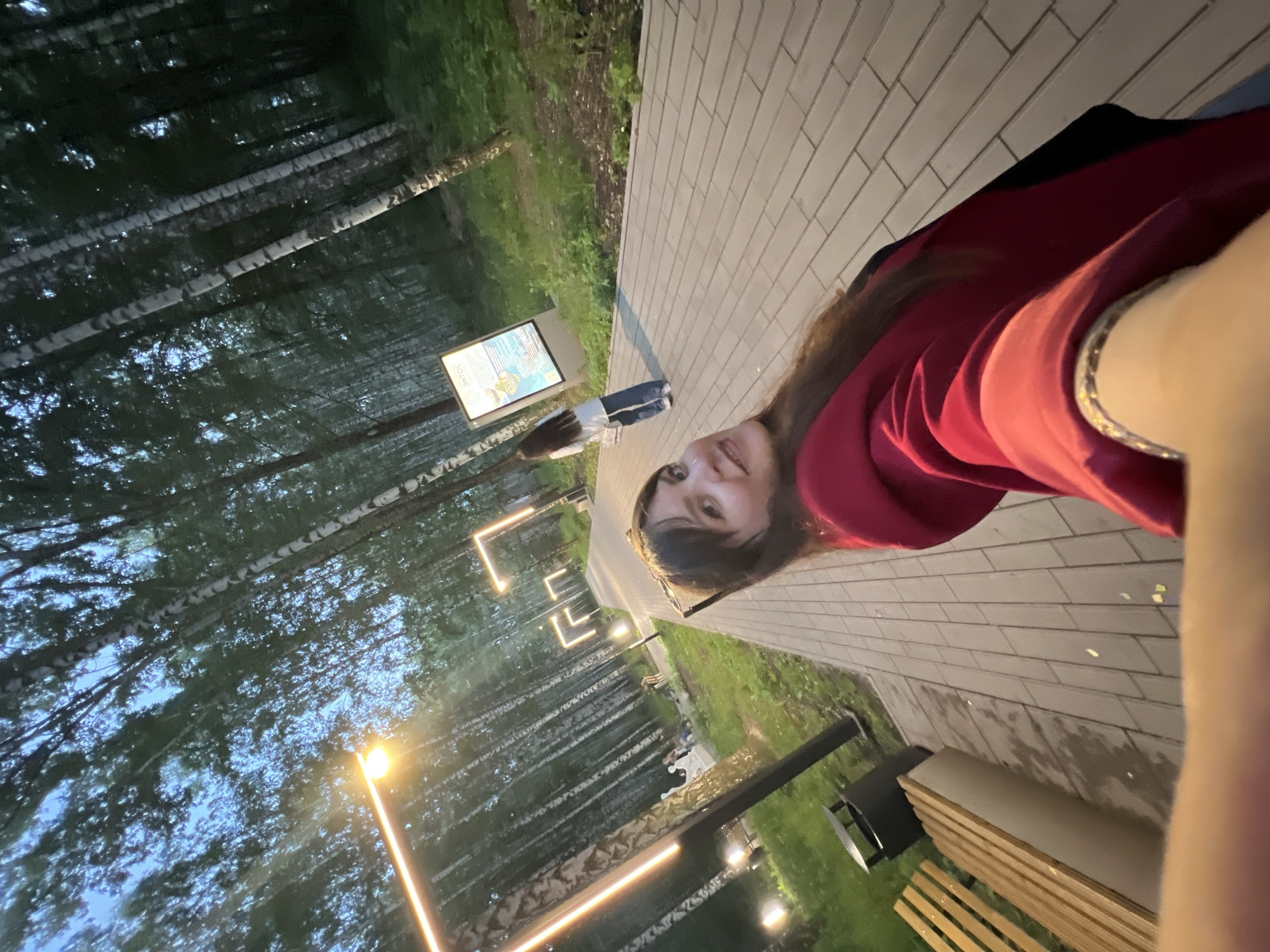 The image depicts a paved walkway in a park lined with birch trees. Modern rectangular light fixtures illuminate the path. In the foreground, a woman in a red hoodie is taking a selfie, smiling at the camera. Further down the walkway, another woman in a white top and jeans is looking at an informational sign. Benches and trash cans are visible along the side of the path. The scene appears to be set in the evening, with the sky a deep blue. The overall environment suggests a public park or recreational area within the city of Chaikovsky, Russia, likely designed for pedestrian use and leisure activities.