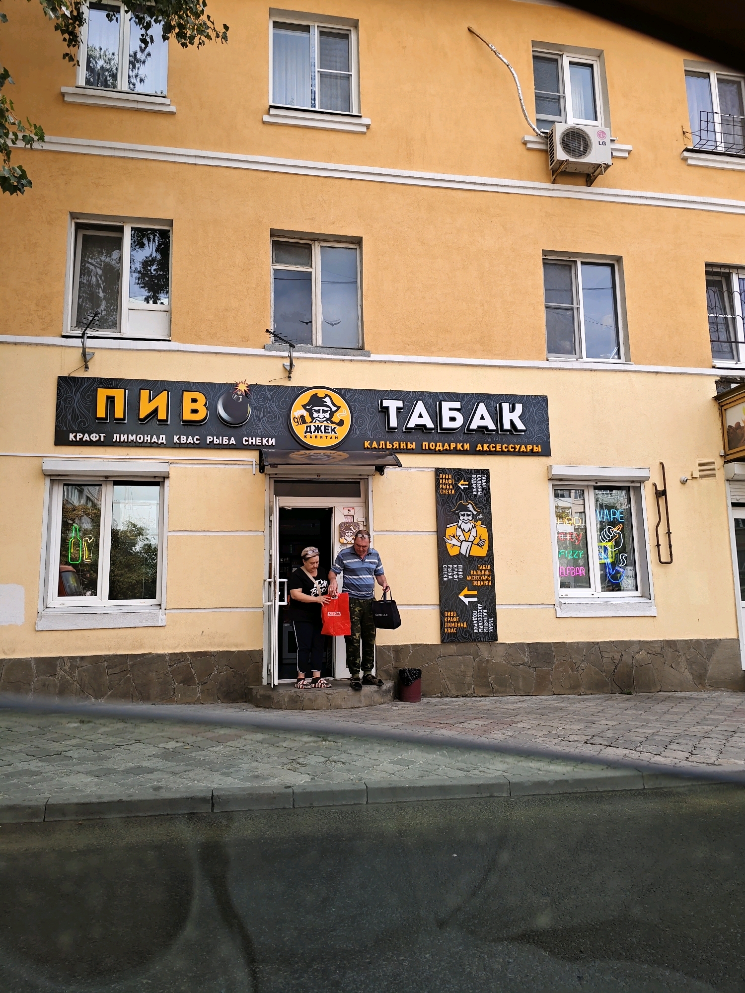 A storefront in Rostov-on-Don, Russia, is captured in this image, featuring a yellow-painted building with multiple windows. A prominent sign above the entrance reads "ПИВО" (Beer) and "ТАБАК" (Tobacco), with smaller text listing "КРАФТ ЛИМОНАД КВАС РЫБА СНЕКИ" (Craft Lemonade Kvass Fish Snacks) and "КАЛЬЯНЫ ПОДАРКИ АКСЕССУАРЫ" (Hookahs Gifts Accessories). Two individuals stand near the entrance, one woman holding a red shopping bag and a man holding a black tote bag. A vertical sign with a pirate-like character and text in Russian is displayed to the left of the entrance. To the right of the entrance, a window displays neon signs advertising "VAPE" and "FIZZY ELFBAB". The building is situated on a street with a paved sidewalk and road. An air conditioning unit is mounted on the exterior wall above the storefront.