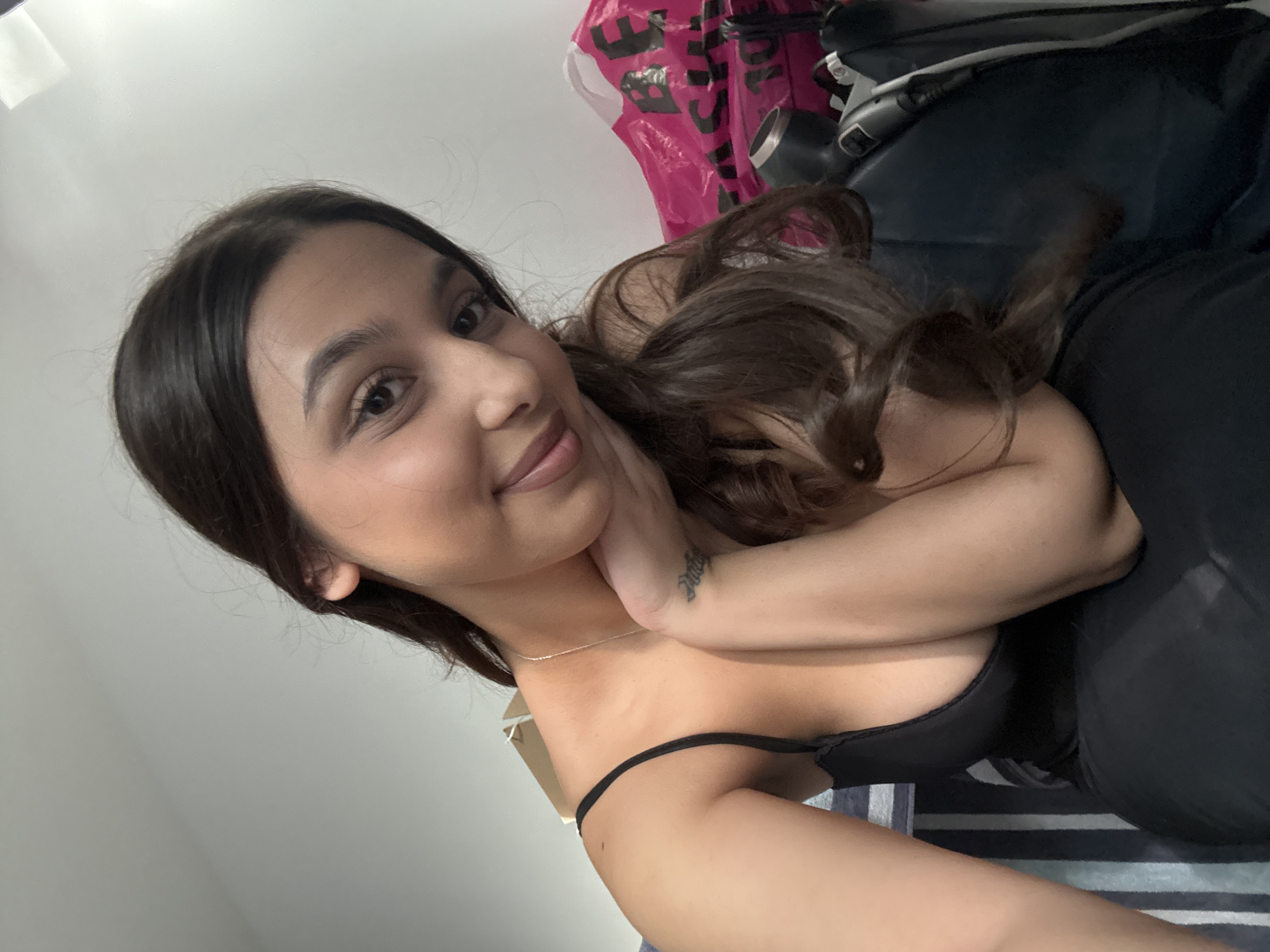 A young woman poses for a selfie indoors. She has dark hair and is wearing a black top. She is smiling slightly. Her right arm is bent and her hand is touching her face. A silver necklace is visible around her neck. A shopping bag with pink lettering is in the background, along with a dark blue object. The background includes a white wall and a striped fabric. There is no information to establish the city as Mönchengladbach, Germany.