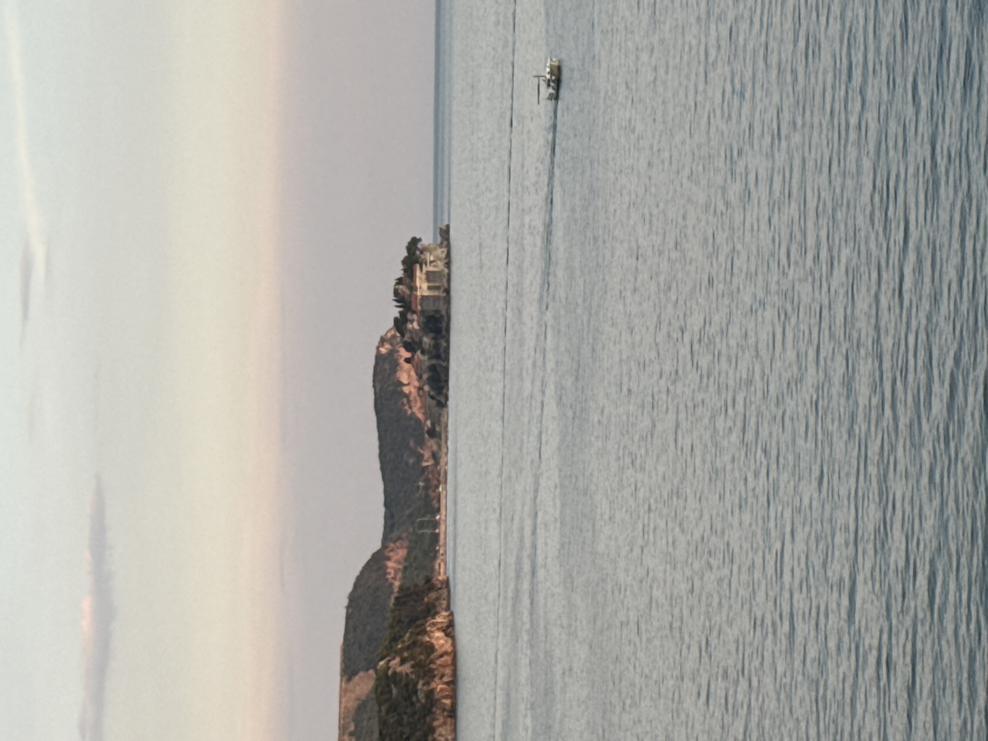 This media file depicts a picturesque scene from Kamenovo, Montenegro, likely captured during the late afternoon or early evening given the soft, diffused light. The image is vertically oriented, presenting a striking contrast between a rugged, tree-covered coastline and the vast, tranquil expanse of the Adriatic Sea.
On the left side, a steep, verdant cliff face dominates the frame. Lush green vegetation clings to its slopes, interspersed with rocky outcrops and hints of buildings nestled into the hillside. The structures appear to be a cluster of houses or a small resort, with warm, earthy tones suggesting terracotta roofs or stone walls, blending harmoniously with the natural landscape. The sheer drop of the cliff down to the water is evident, adding a dramatic element to the scene.
To the right, the sea stretches out, its surface rippled by gentle waves, reflecting the muted tones of the sky. The water is a cool, grayish-blue, creating a sense of peace and serenity. A small, white motorboat is visible in the distance, its wake trailing behind it as it navigates the calm waters. The boat is a solitary object, emphasizing the scale and openness of the seascape.
The sky above is a soft, pale gradient, transitioning from a hazy white or very light gray near the top to warmer, peachy hues closer to the horizon, hinting at the approaching sunset. There are no visible clouds, suggesting clear weather. The overall impression is one of natural beauty and tranquility, with a touch of human habitation integrated into the stunning coastal environment. No visible text is present in the media file.