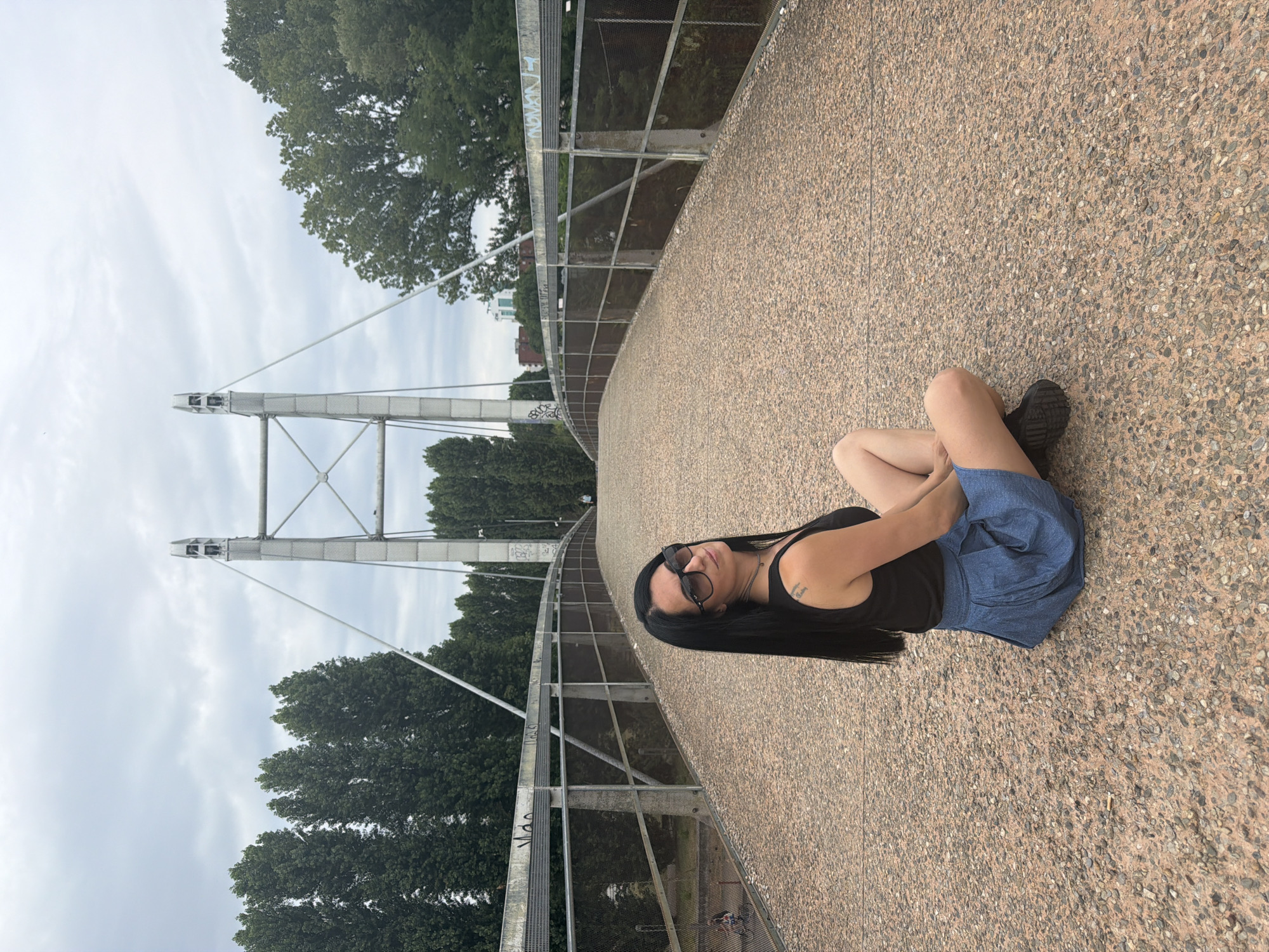 A woman with long black hair and sunglasses is sitting cross-legged on a pedestrian bridge in Corsico, Italy. She is wearing a black tank top and a blue denim skirt. The bridge has a metal railing and is paved with small stones. In the background, there are tall trees and a cloudy sky. The bridge appears to lead to a modern suspension structure.