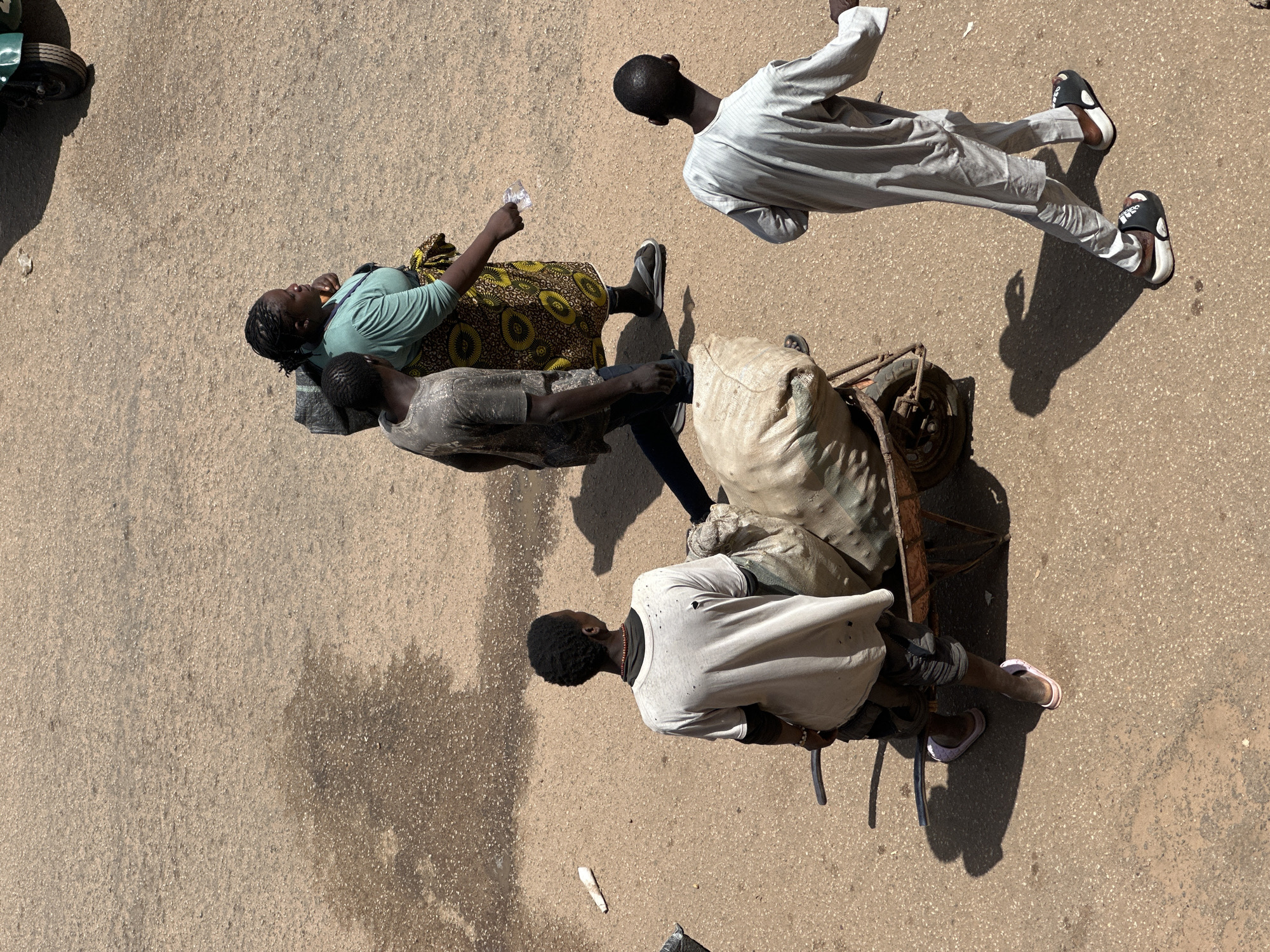 In Bukuru, Nigeria, four individuals traverse a paved street under a bright sun, casting long shadows. In the foreground, a young male, wearing a loose-fitting light-colored shirt and dark pants, pushes a two-wheeled cart loaded with two large, tan sacks. He wears sandals. To the right, another young male walks away from the viewer, dressed in a light-colored, long-sleeved tunic and trousers, also wearing sandals. Further in the background, a woman in a turquoise top and a patterned skirt walks with a young male in a grey t-shirt and dark pants. The woman holds a small, clear object in her hand. The environment appears to be an outdoor market or street scene, with the ground a textured, light brown asphalt. A portion of a green vehicle is visible in the upper right corner.