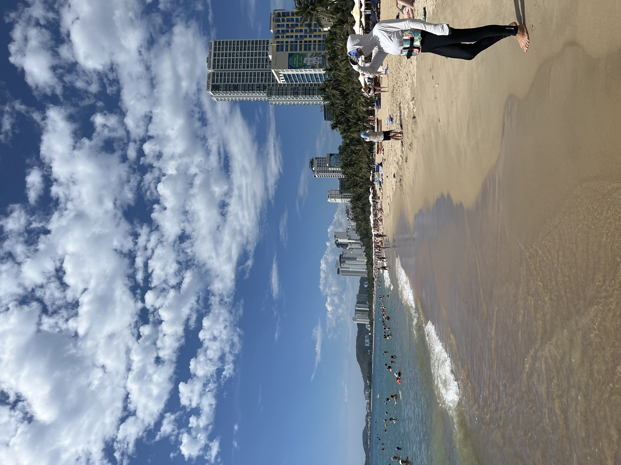 The image depicts a crowded beach scene in Nha Trang, Vietnam, likely during daylight hours given the bright sun and clear sky with scattered white clouds. In the foreground on the right, a person is standing on the sandy shore, wearing a white long-sleeved hooded top, black leggings, and a colorful life vest or flotation device. Their face is partially obscured as they appear to be shielding their eyes from the sun.
The beach itself is a wide expanse of light brown sand, leading to the ocean where numerous people are swimming, wading, and enjoying the water. The water is a clear blue-green, with small waves breaking near the shore. Along the coastline, beyond the beach and the water, a line of palm trees and modern high-rise buildings are visible, indicative of a developed urban area adjacent to the coast. The architecture of the buildings suggests a tourist-oriented city. The overall atmosphere is one of a lively beach resort.
