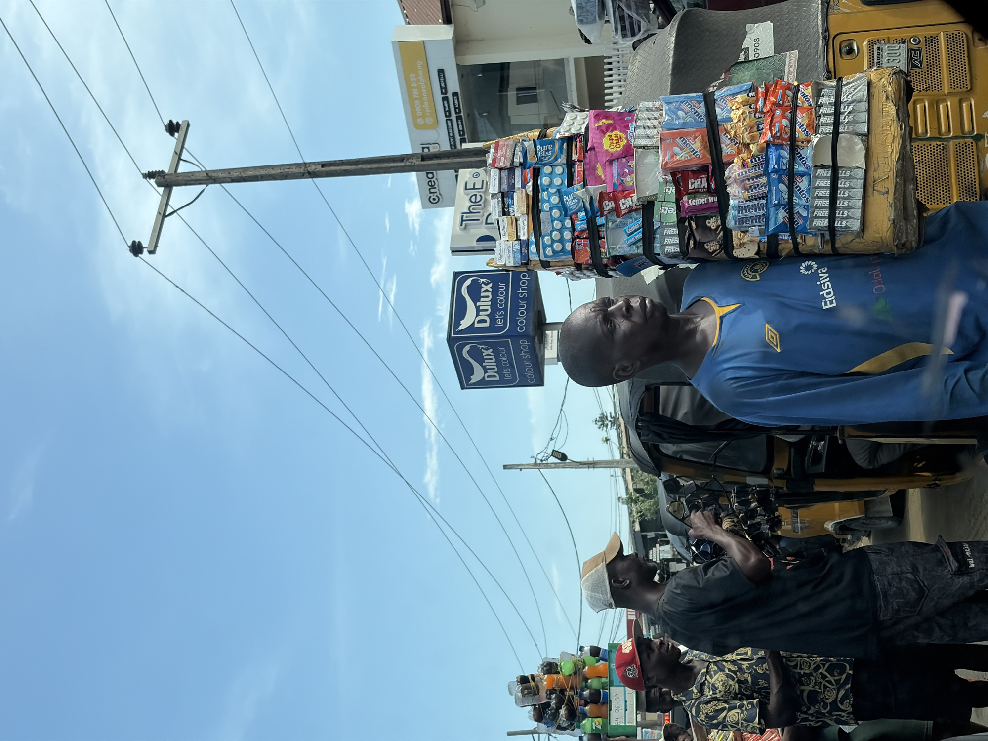 The image depicts a bustling street scene in what appears to be a commercial area in Ado, Nigeria, under a bright blue sky with sparse clouds, indicating daytime. The primary activity is street vending, with several individuals gathered around makeshift stalls.
In the foreground, a man with a bald head wears a blue and yellow soccer jersey, featuring "Eidsiva" and "Odal p" sponsor logos. He stands beside a yellow motorized tricycle, resembling a Keke Napep, which serves as a mobile vending stall. The tricycle is heavily laden with various packaged goods, including "Mentos" mints (in multiple flavors), "Chupa Chups" lollipops, "Pure Bliss" biscuits, "Rothmans" cigarettes, "Center Tru" candies, "CRA" snacks, and items labeled "FREE LLS," all secured with black straps.
Further back, another man in a black t-shirt and a light-colored cap is visible, facing away from the camera, near another tricycle. A third man wearing a patterned shirt and a red cap is partially seen to the right. Another small stall with various bottles stacked on top is present in the mid-ground.
Street infrastructure includes numerous wooden utility poles with multiple power lines traversing the sky. In the background, commercial buildings are visible. Several signs display prominent text: large "Dulux let's colour colour shop" signs, a yellow sign for "ryde.onealpha.ng" with the phone number "0908 781 8152" and locations listed as "PORT HARCOURT WARRI UGHELLI BENIN AGABOR." Other partial signs include "The E" and "Cnea." Text on the yellow tricycle includes "ACO," "1300," and "0908." The man in the black t-shirt has "balencia" written on his pants. The scene conveys a sense of everyday commerce and urban activity.