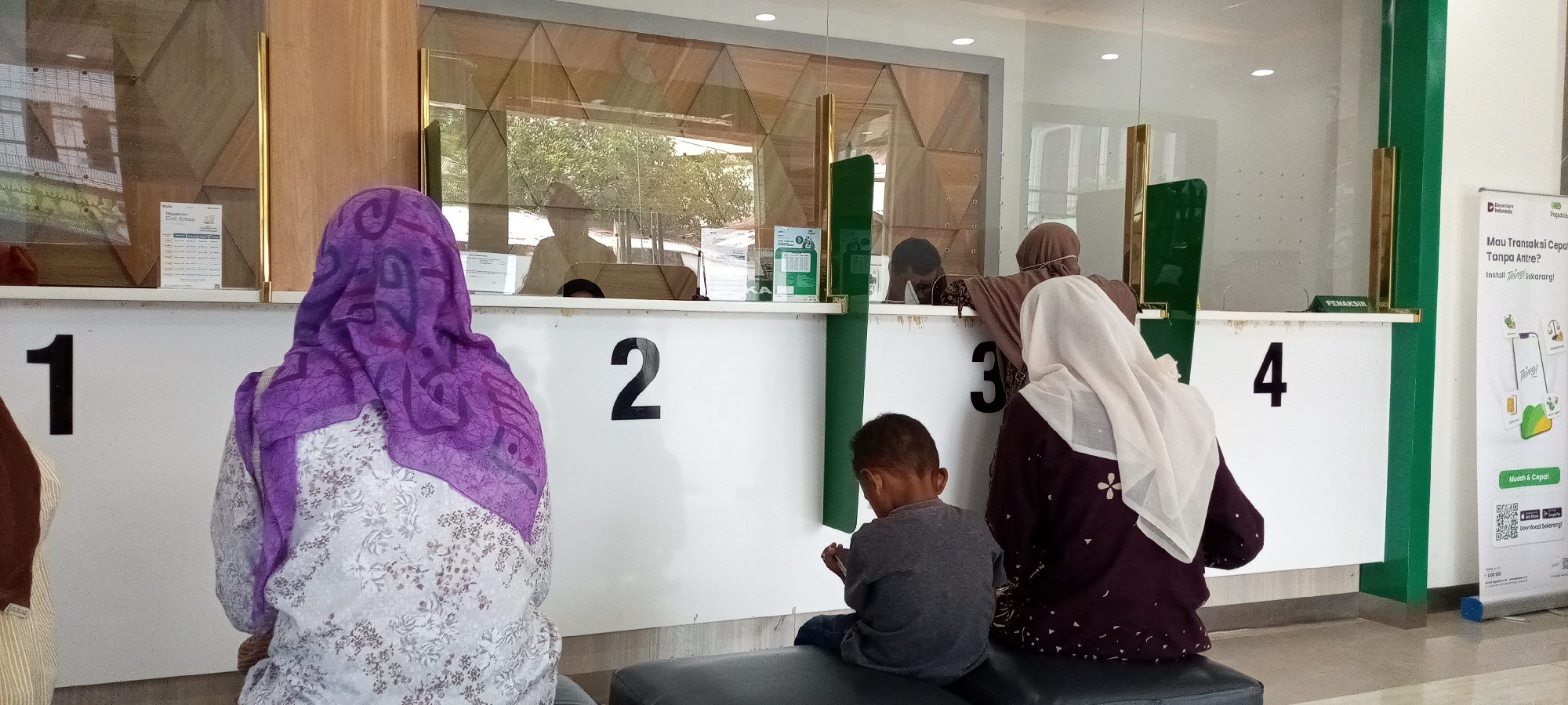 The image depicts an interior scene, likely a bank or government office, with a numbered counter system. There are four visible counter numbers: 1, 2, 3, and 4. In the foreground, from left to right, are three individuals waiting. The first is a person wearing a purple and white patterned hijab and a light-colored patterned top, seen from behind. To their right, a young child wearing a gray t-shirt and blue jeans sits on a bench, facing away from the camera. Next to the child is a person wearing a light cream-colored hijab and a dark maroon top with white floral patterns, also seen from behind. Behind the counter, through a glass partition, at least two individuals are visible, appearing to be employees interacting with customers. One employee, wearing a dark hijab, is seen leaning forward. The environment appears to be a waiting area with a clean, modern aesthetic, featuring wooden accents and green vertical dividers. A promotional banner with Indonesian text is visible on the far right, advertising a mobile application. The overall impression is a public service space where people are waiting for their turn at the counter.
