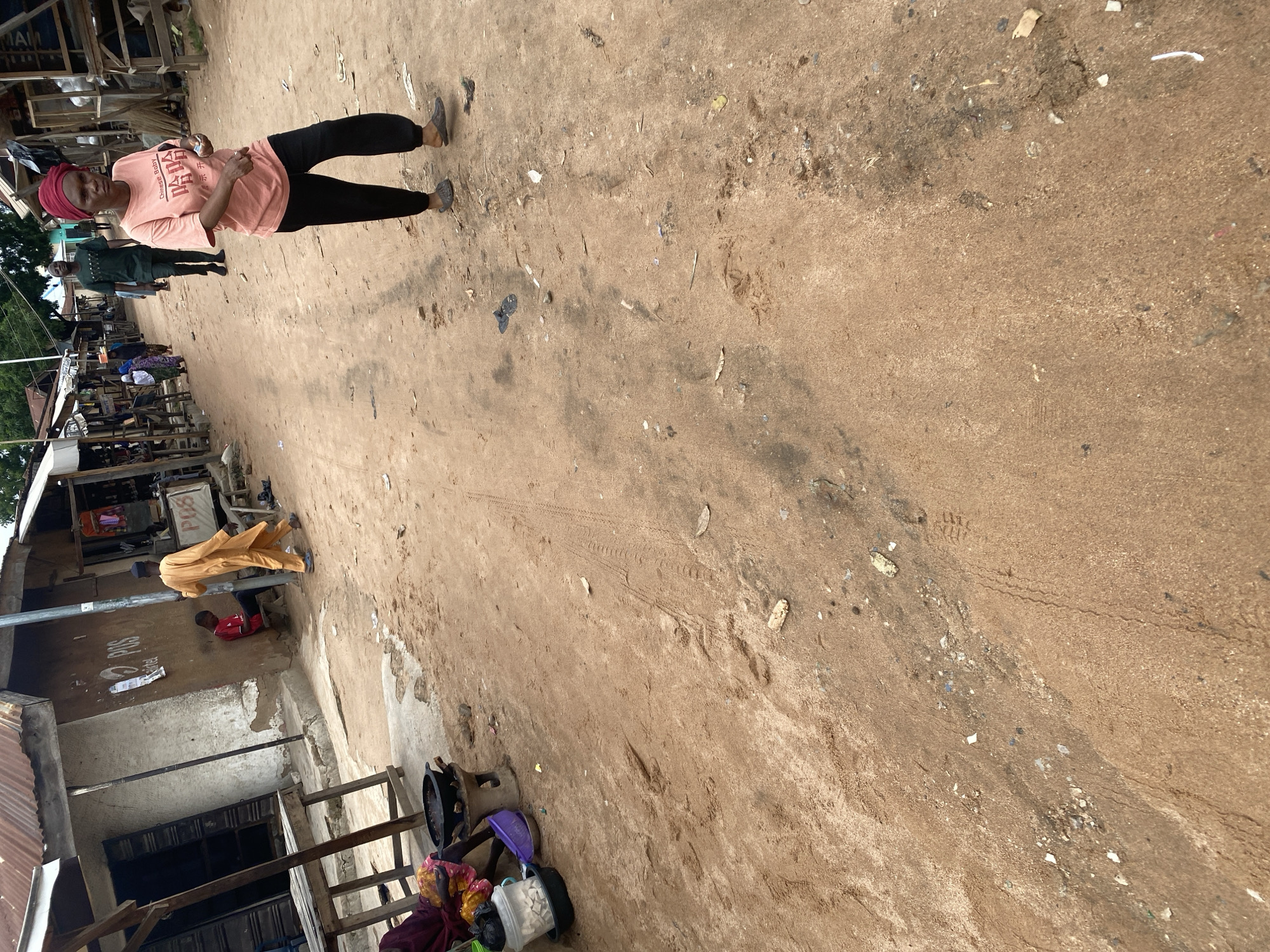 In an outdoor marketplace scene in Jalingo, Nigeria, a woman wearing a red headscarf and a pink t-shirt stands in the center, holding a small white object. She is looking towards the viewer with a neutral expression. To her left, a person in a dark green outfit walks away from the camera. Further in the background, stalls with various goods are visible. In the foreground and to the right, the ground is covered in dry, sandy dirt, with some debris and tire tracks.
On the left side of the image, a man in a yellow garment bends down, interacting with a young boy in a red shirt who is seated. Behind them, a rustic structure with a corrugated iron roof and wooden elements suggests a simple marketplace setting. The overall impression is one of a bustling, everyday scene in a somewhat dusty environment. The lighting suggests it is daytime.