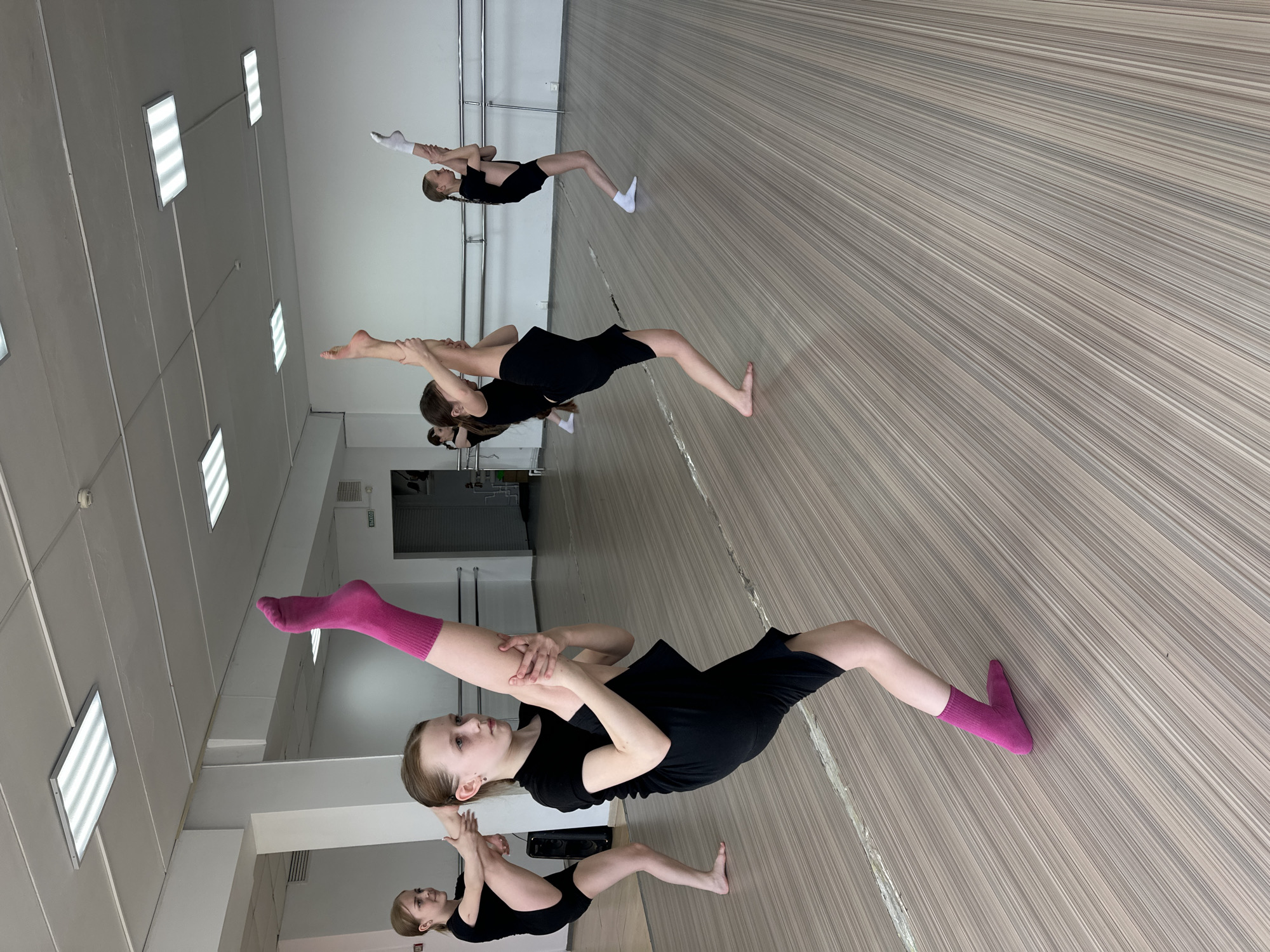 The image shows a dance studio in what seems to be Votkinsk, Russia. Several young dancers dressed in black leotards and shorts are stretching. The studio has a light-colored wood floor, white walls, and fluorescent lights on the ceiling. Some dancers have socks on their feet, while others are barefoot. The dancers are holding one leg up towards their head, stretching their hamstrings. The scene is well-lit and appears to be during a dance class or practice session.