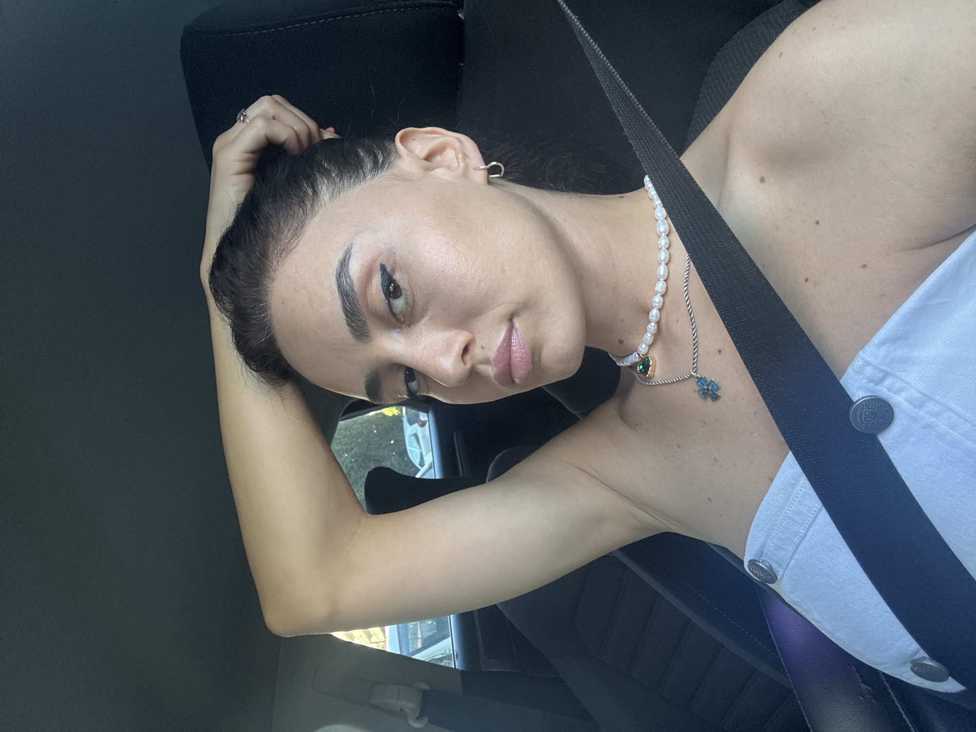 A portrait of a young woman sitting in a car, looking to the right with her head tilted back. She has dark hair pulled back, her left arm is raised and her hand is resting on her head. She is wearing a white top, a pearl necklace with a green pendant, and a silver hoop earring in her left ear. A black seatbelt is visible across her chest. The setting appears to be daytime, with sunlight filtering into the car. The background shows the interior of the car, including the seat and part of the dashboard. There are no visible text elements in the image. The woman's expression is neutral, with a slight hint of a smile. The overall mood is relaxed and casual.