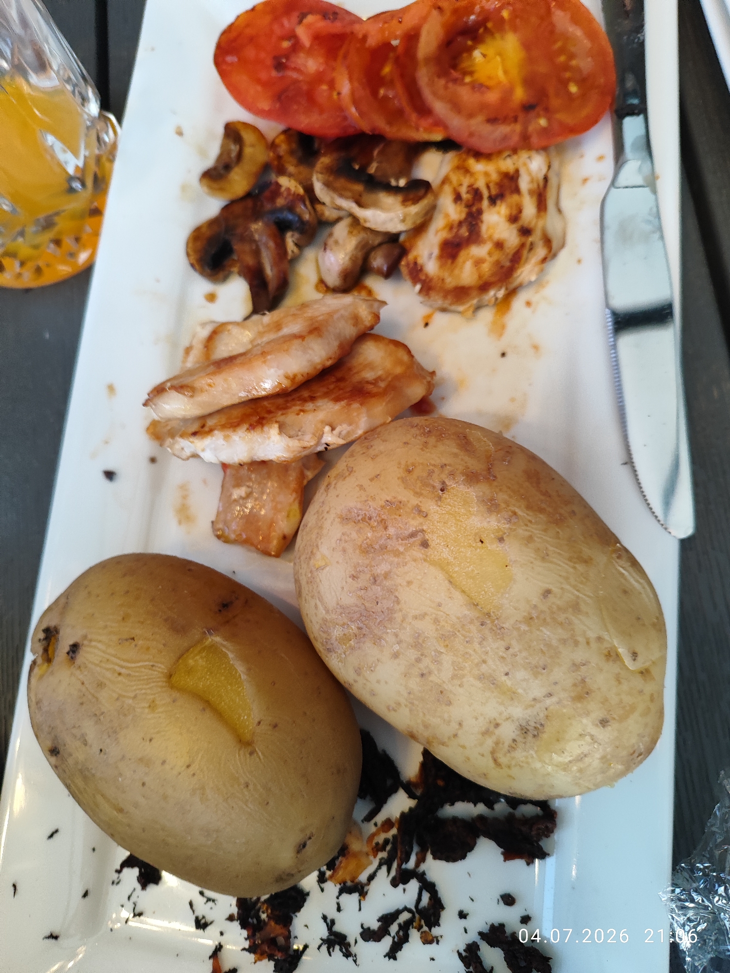 This is a close-up, high-angle shot of a white rectangular plate filled with food. In the foreground, there are two whole baked potatoes. The potato on the left has a visible yellow interior where it appears to have been cut or cracked open. The potato on the right is also whole. The plate contains other food items, including sliced tomatoes, grilled mushrooms, and pieces of cooked chicken. A portion of a glass containing an orange beverage is visible in the upper left corner. A silver knife with a serrated edge rests on the right side of the plate, next to the food. The background is dark and out of focus. There is no discernible location within Wellingborough, United Kingdom, or any indication of people, actions, or interactions. The timestamp "04.07.2026 21:06" is superimposed on the bottom right of the image.
