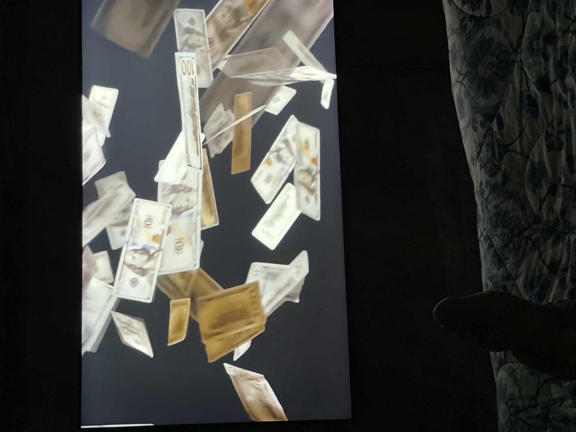 The image shows a close-up of a television screen displaying falling US hundred-dollar bills. In the foreground, a person's foot is visible, resting on a patterned blanket. The scene appears to be indoors, with the television screen taking up the majority of the frame. The lighting is dim, highlighting the illuminated screen. There is no discernible connection to Pattaya City, Thailand in this image.