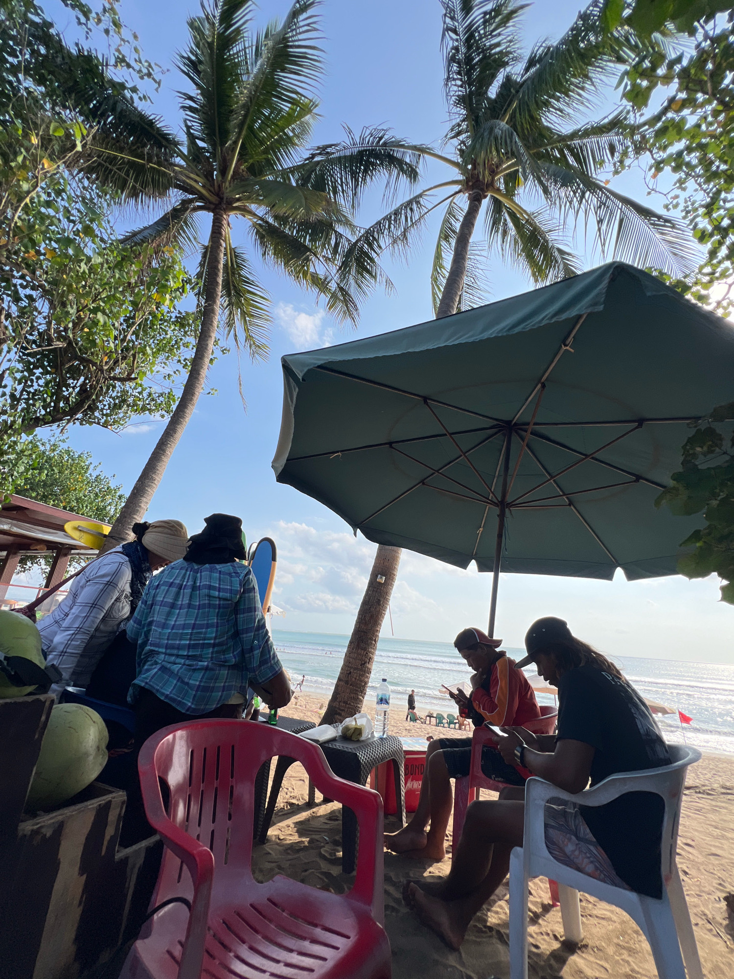 The image depicts a beach scene in Kuta, Indonesia. The foreground features several people gathered under a large green umbrella. Two individuals are seated on chairs, engrossed in their mobile phones. To the left, there are two more people standing and looking towards the ocean. Several large green coconuts are visible, suggesting a refreshment stand or a local vendor. The background reveals a sandy beach with calm ocean waters and a clear blue sky with scattered clouds. Palm trees line the edge of the beach, casting shadows on the sand. The overall atmosphere appears relaxed and casual, indicative of a typical day at this popular tourist destination.