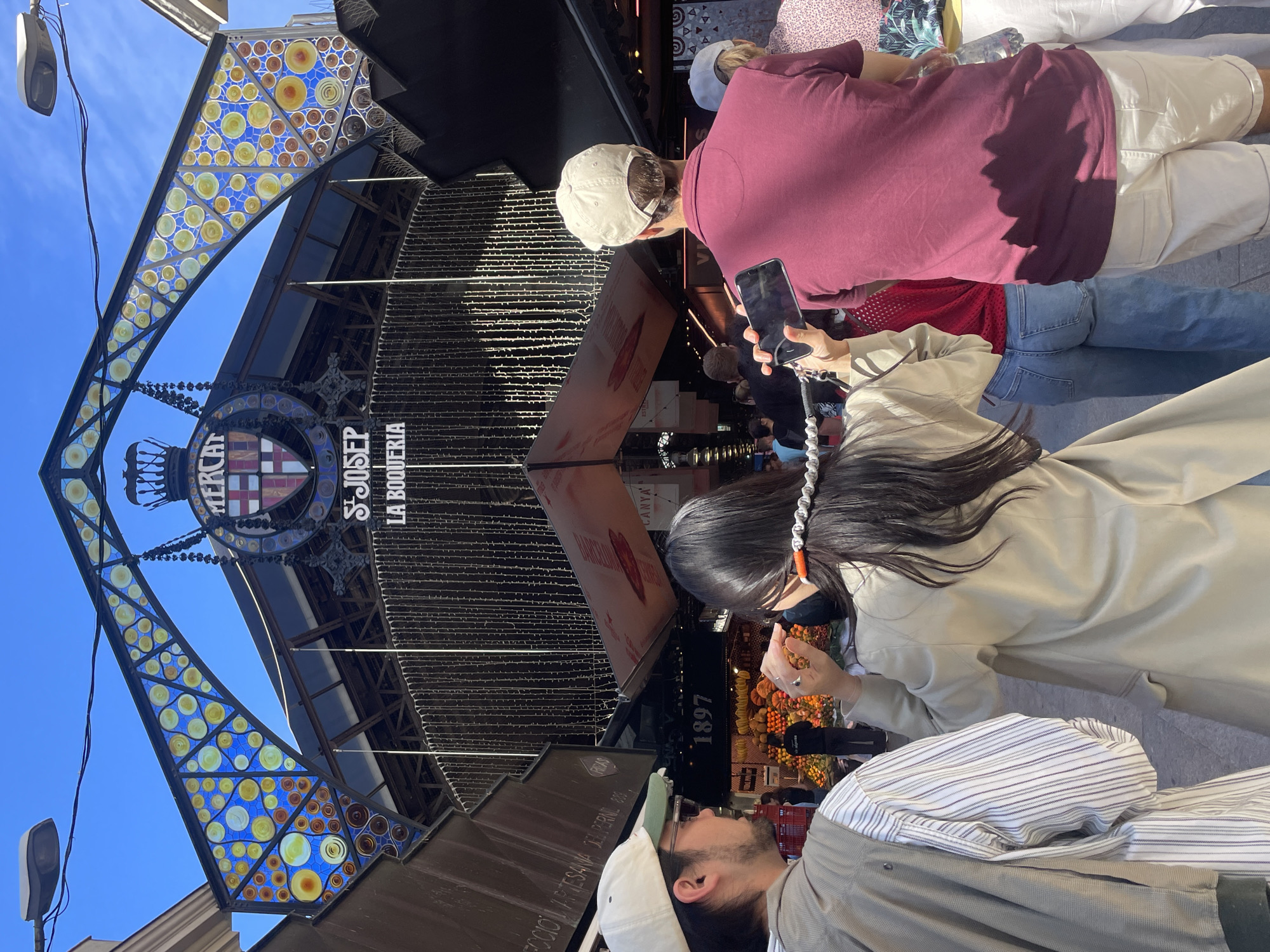 The image shows a group of people at the entrance of La Boqueria market in Barcelona, Spain. The market's signage, including the Mercat de Sant Josep La Boqueria name and 1897 date, is visible. The entrance features colorful stained glass details and hanging lights. People are standing and looking, and some are holding and using mobile phones.
