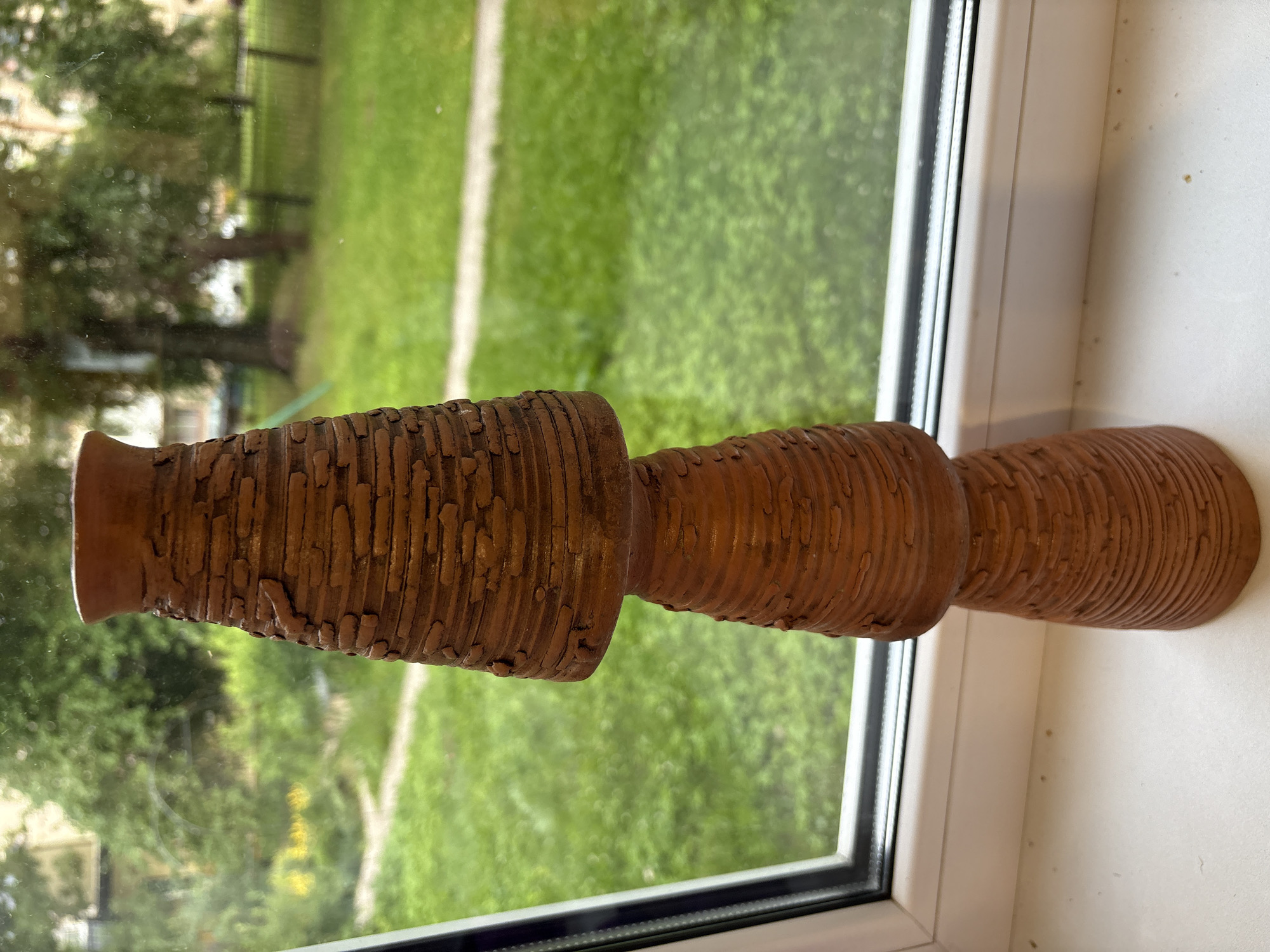 A stacked, terra cotta vase with a textured, horizontal ribbed pattern sits on a white windowsill. The vase is light brown and tapers in sections, with the top section being the widest. Behind the vase is a window that looks out onto a blurred green lawn and trees. The scene appears to be outdoors during the daytime.