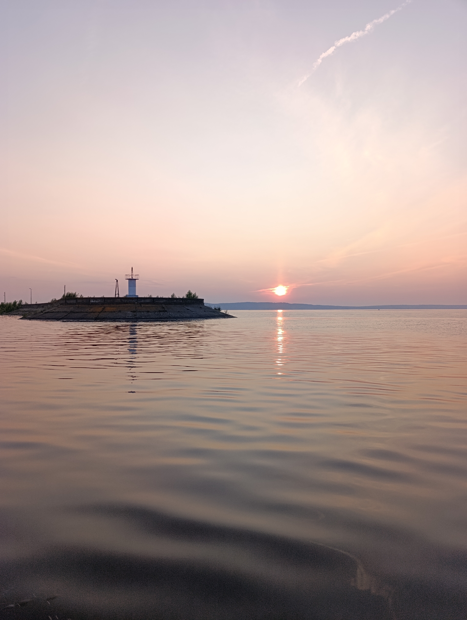 This image depicts a serene sunset over the Kama River in Chaikovsky, Russia. In the foreground, the calm water reflects the soft hues of the sky, with gentle ripples creating a textured surface. To the left, a man-made structure, possibly a pier or breakwater, juts out into the river. A white lighthouse with a visible lantern room stands prominently on this structure. The structure itself is made of concrete, with a low wall bordering its perimeter. Trees and some vegetation can be seen on the raised platform. In the distance, the sun is setting, casting a warm orange glow across the horizon and its reflection shimmers on the water. A faint cloud trail stretches across the pale pink and lavender sky. The overall scene is tranquil and picturesque, capturing the beauty of a twilight moment on the river.