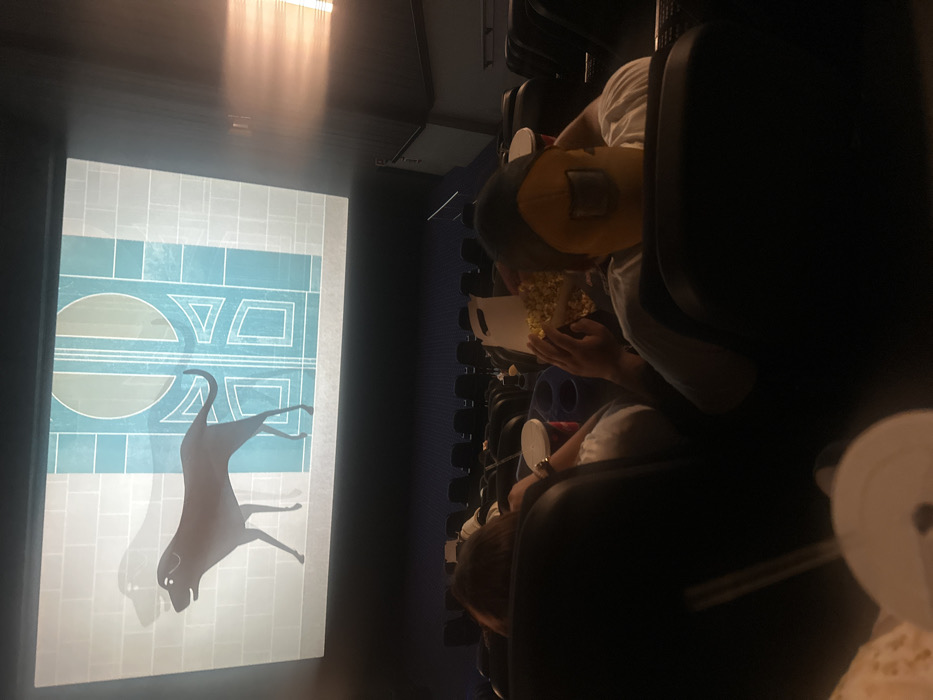 The image depicts the interior of a darkened movie theater in Mexico City, Mexico, viewed from an elevated perspective behind seated patrons. A large projection screen dominates the upper frame, displaying an animated scene. The scene features a stylized, dark-colored dog, resembling a Xoloitzcuintli (likely from Disney/Pixar's *Coco*), walking left against a light grey wall with a faint dog shadow, and an ornate teal-colored door with geometric patterns.
In the midground, two individuals are visible from behind, seated in dark, upholstered theater chairs. The person on the left, with brown hair, wears a white shirt; a red cup with a white lid and a white-lidded cup with a straw are in cup holders to their right. The person on the right, wearing a white t-shirt and a distinct yellow and black cap (resembling a welding mask design), holds a white cardboard tub filled with popcorn with both hands, actively consuming it. Rows of dark blue or black empty seats extend into the background beyond these individuals. The right side of the auditorium features a wall with a prominent horizontal light flare emanating from an unseen source.