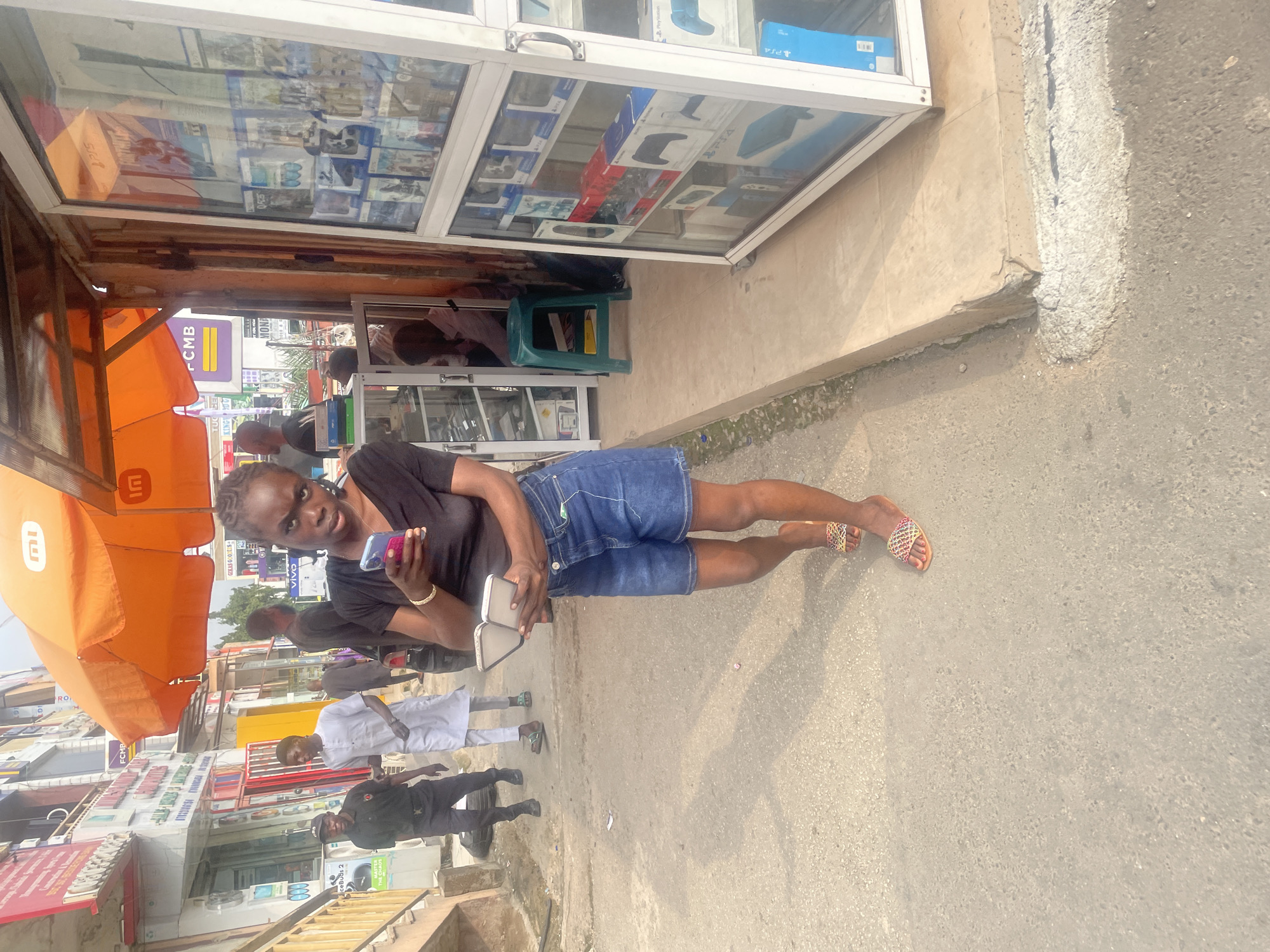 The image depicts an outdoor scene on a paved street, likely in Abuja, Nigeria, with several people and a commercial stall. In the foreground, a young woman is looking down at a mobile phone in her hands. She is wearing a black t-shirt and denim shorts, and colorful sandals. Behind her, a security guard in a black uniform and a man in a white traditional outfit are walking away from the camera.
To the right of the frame is a retail stall with a glass display case showcasing electronic devices, including what appear to be video game consoles and accessories. An orange awning extends over the stall. In the background, other people are visible, along with buildings and signage, suggesting a busy urban environment. The ground is a mix of asphalt and concrete. The overall lighting suggests it is daytime.