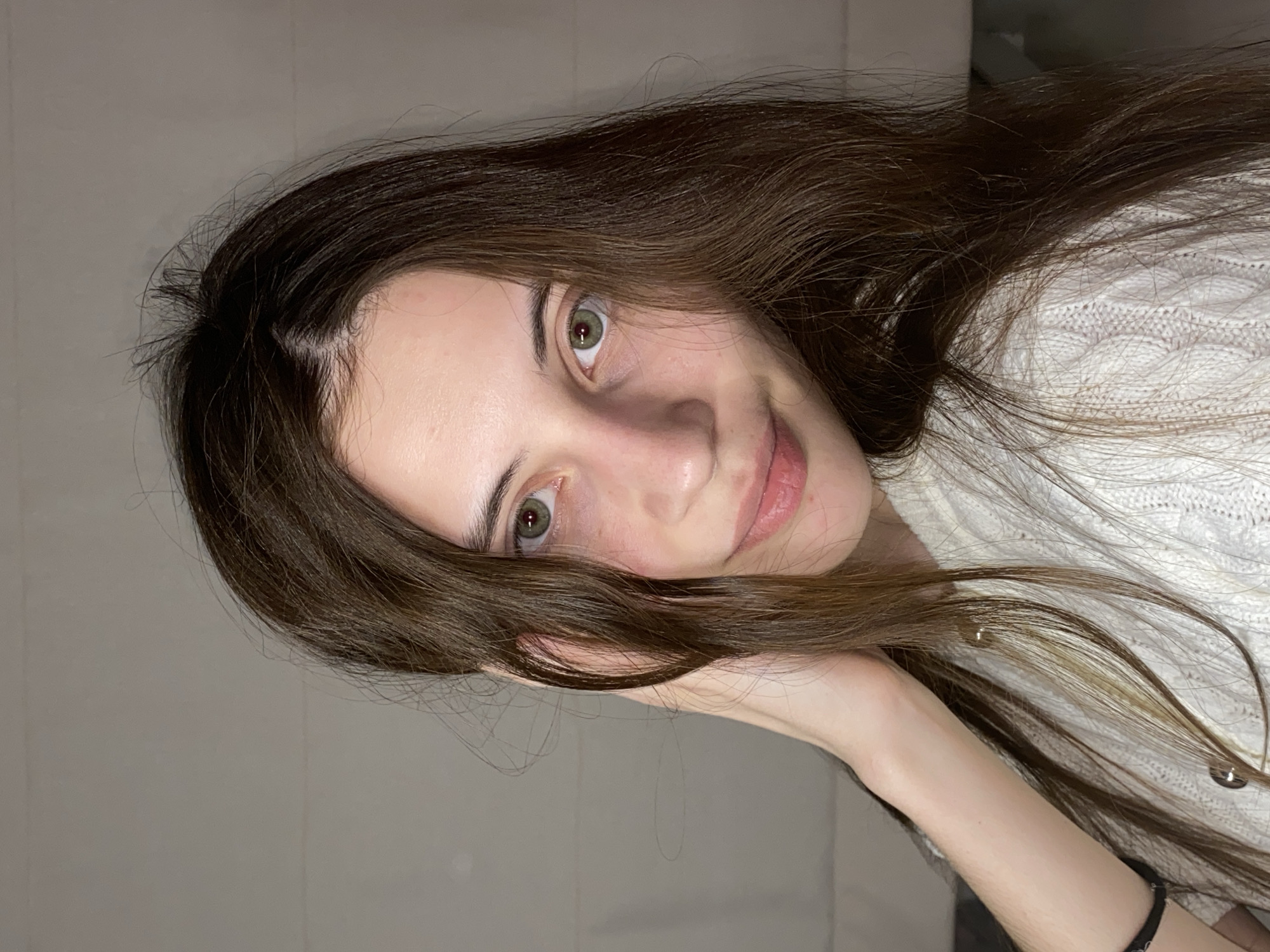 The image is a close-up portrait of a young woman with long, dark brown hair, presenting a gentle expression and a slight smile while looking directly at the camera. Her hair, appearing slightly wavy and natural, is parted on her left side and flows around her face and over her right arm. She has light skin, well-defined dark eyebrows, and striking green/hazel eyes that show intricate details within the iris. Her lips are a natural pink tone and are softly parted in a subtle smile. She is wearing a white, cable-knit sweater, partially visible on her right side. Her right hand or arm is seen, seemingly supporting her head or resting beneath her hair, and a black hair tie is visible on her wrist. Notably, there are a few distinct, spherical reflections on strands of her hair that resemble water droplets. The background is a plain, light gray or beige wall, indicating an indoor setting with soft, even lighting. The photograph was taken in Votkinsk, Russia. There is no visible text in the image.