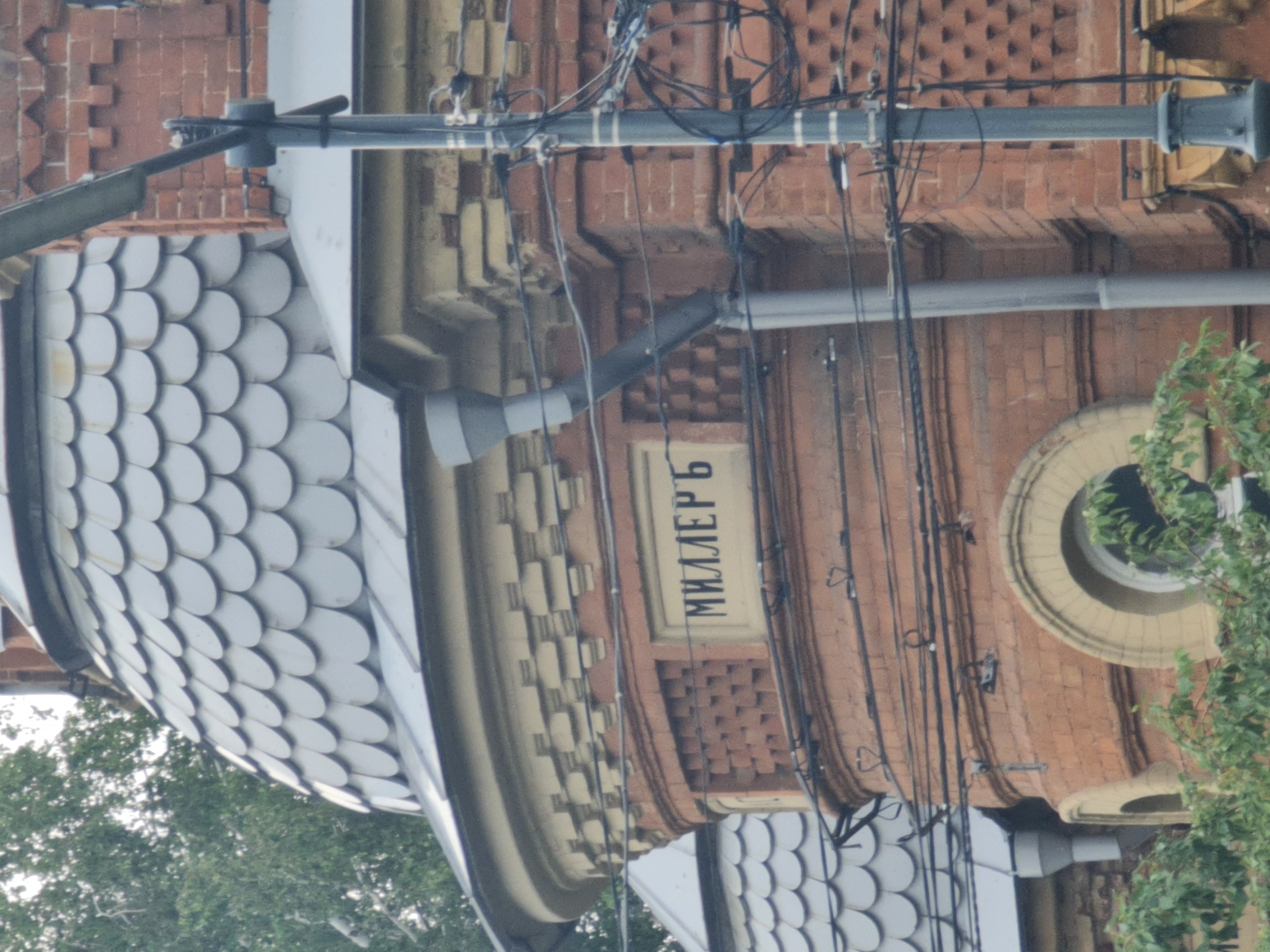 The image is a close-up, eye-level shot of a brick building with a domed roof and some architectural details. On the curved brick surface, a rectangular plaque with Cyrillic lettering is visible. The lettering appears to read "МИЛЛЕРЪ." The building is partially obscured by trees and a lamppost with numerous wires. The architectural style suggests it is an older building, likely from the late 19th or early 20th century. The building is located on Ulitsa Karla Marksa in Irkutsk, Russia, and is part of the historical architecture of the city.