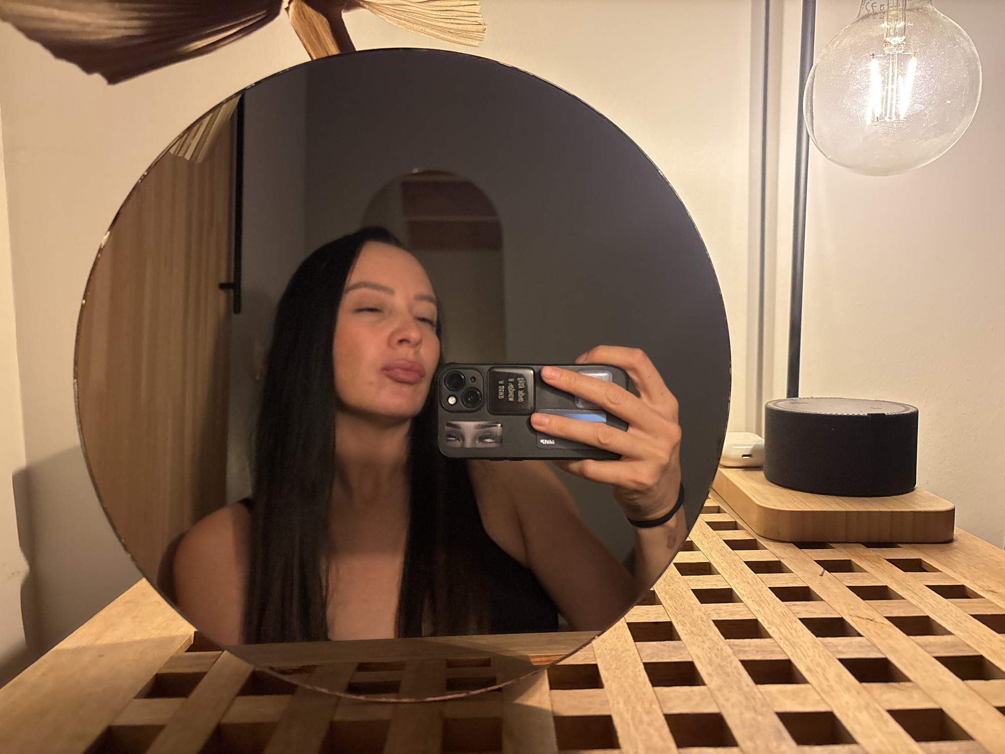 A woman is taking a selfie in a round mirror. She has long dark hair and is sticking her tongue out. She is wearing a black top. The phone she is using has a black case with stickers on it. The mirror is sitting on a wooden lattice table. There is a black cylindrical object and a lightbulb in the background. The location is not evident from the image.