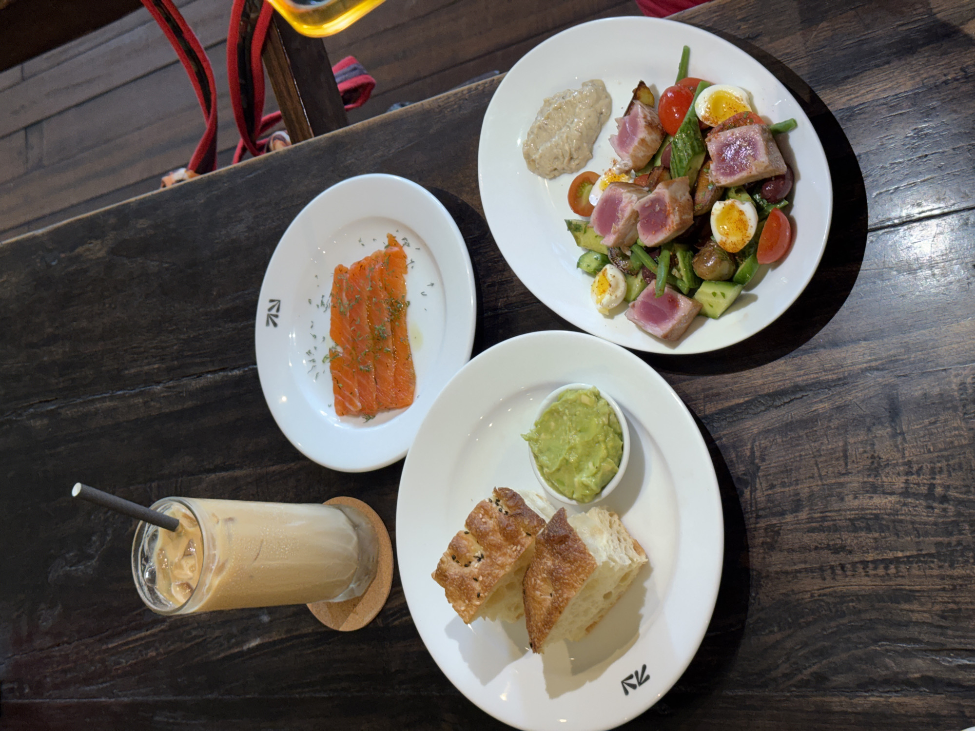 The image shows a table at a restaurant, likely in North Kuta, Indonesia. A wooden table is set with four plates. One plate holds sliced salmon. A second plate has a tuna salad with avocado, green beans, tomatoes, and boiled eggs, along with a scoop of dip. The third plate holds slices of bread and guacamole. A tall glass of iced coffee with a straw sits on a coaster next to the plate of bread and guacamole. Some red straps are visible in the background, possibly belonging to a backpack.