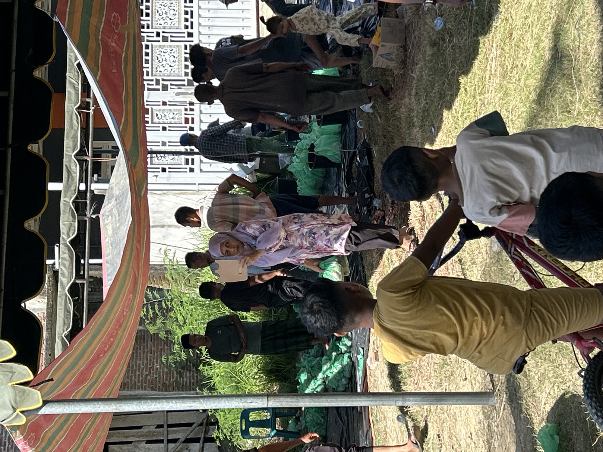A daytime outdoor scene in Lhokseumawe, Indonesia, presents a group of people gathered under a striped tent structure, possibly for a community event or meeting. The setting includes green vegetation and a partial brick wall. Several individuals are standing or sitting, some of whom are interacting with others. One woman wearing a light purple floral hijab is holding a white document. A boy in a yellow shirt sits on a bicycle. Other figures are dispersed throughout the scene.