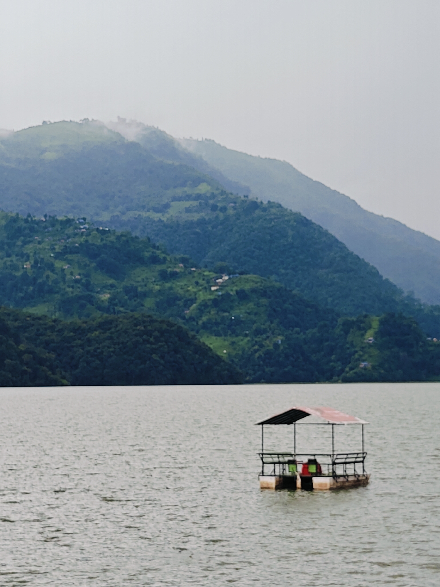 The image depicts a serene natural landscape on a body of water, likely Phewa Lake, in Pokhara, Nepal. In the foreground, a small, flat-bottomed floating structure, possibly a raft or a small boat with a canopy, is visible in the center of the frame. The structure has a dark frame and a reddish-brown canopy, and appears to be equipped with seating. Two individuals are seated on the structure, one in green and another in red. The water is calm with a slightly rippled surface, reflecting the overcast sky. In the background, lush green hills rise dramatically, covered in dense vegetation and dotted with small scattered buildings, indicating a rural or semi-rural settlement on the slopes. The hills recede into the distance, with the farthest peaks shrouded in a soft, hazy mist, creating a sense of depth and atmosphere. The overall scene conveys a peaceful and picturesque view of a lakeside setting with mountainous terrain.