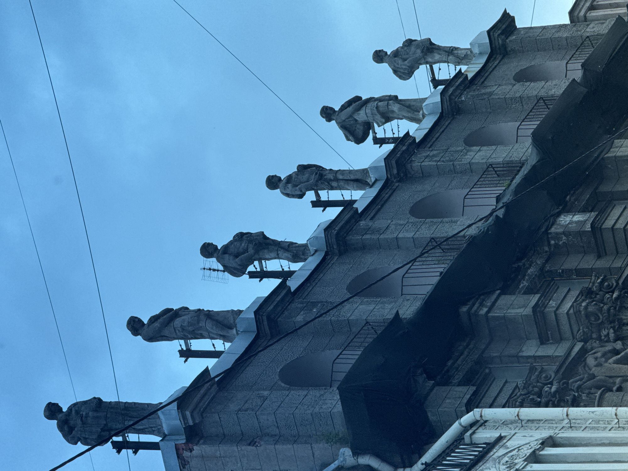 The image shows a row of six stone statues of men standing atop a building in Saint Petersburg, Russia. The statues are positioned along the roofline of the building, which appears to be made of grey stone. The sky behind the statues is a deep blue, suggesting it is either dusk or dawn. The building itself has several arched windows and balconies, and some electrical wires are visible strung across the sky. The overall scene is one of architectural grandeur, with the statues adding a sense of historical significance and artistic expression to the urban landscape. The statues are weathered and appear to be quite old, contributing to the atmosphere of history and timelessness.