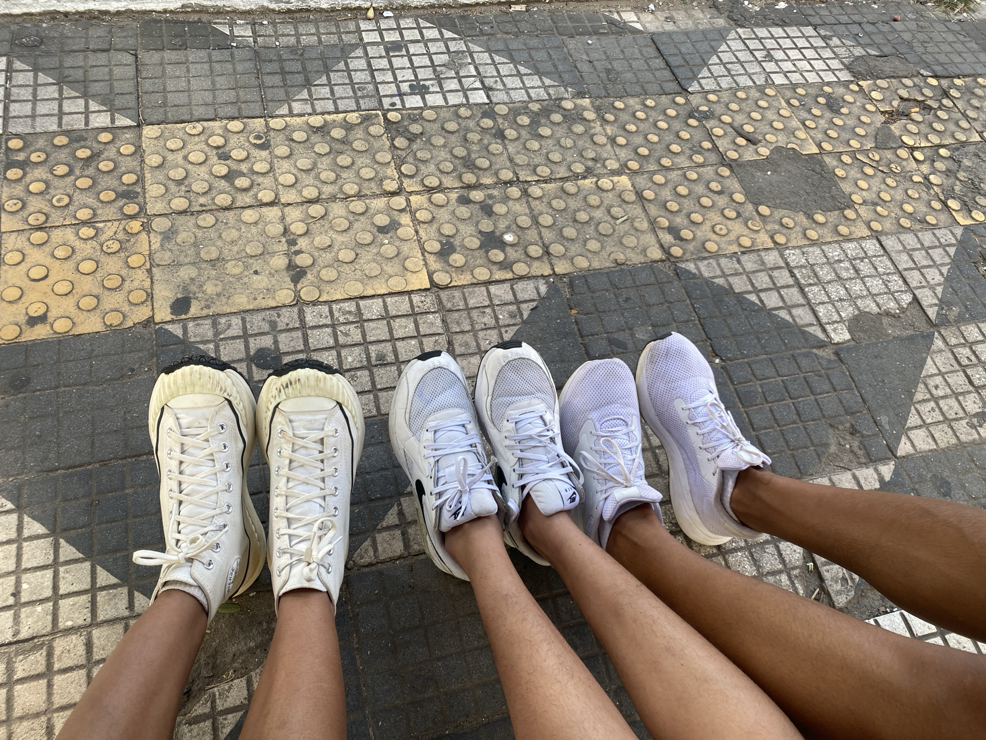 The image shows three pairs of feet wearing white sneakers on a patterned sidewalk. From left to right: the first pair of feet are wearing high-top canvas sneakers with a thick, textured sole; the second pair are wearing white mesh running shoes with black Nike logos; the third pair are wearing white running shoes with a slight lavender tint. The sidewalk is made of square tiles in a alternating checkerboard pattern of gray and white squares, and yellow squares with circular bumps. The photograph seems to be taken at a pedestrian sidewalk, on the city of São Paulo, Brazil.