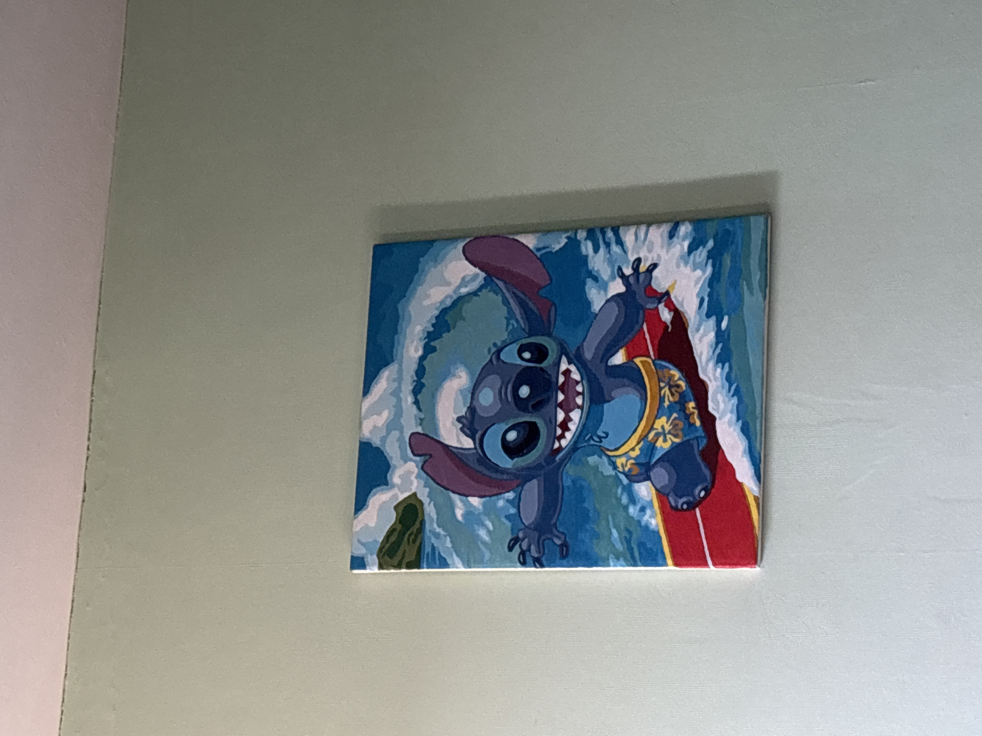 A painting of Stitch from the Disney movie "Lilo & Stitch" hangs on a light green wall. Stitch is depicted surfing, wearing a yellow Hawaiian shirt. The background is blue and white, representing waves. The painting is rectangular. The location is Novoe Devyatkino, Russia.