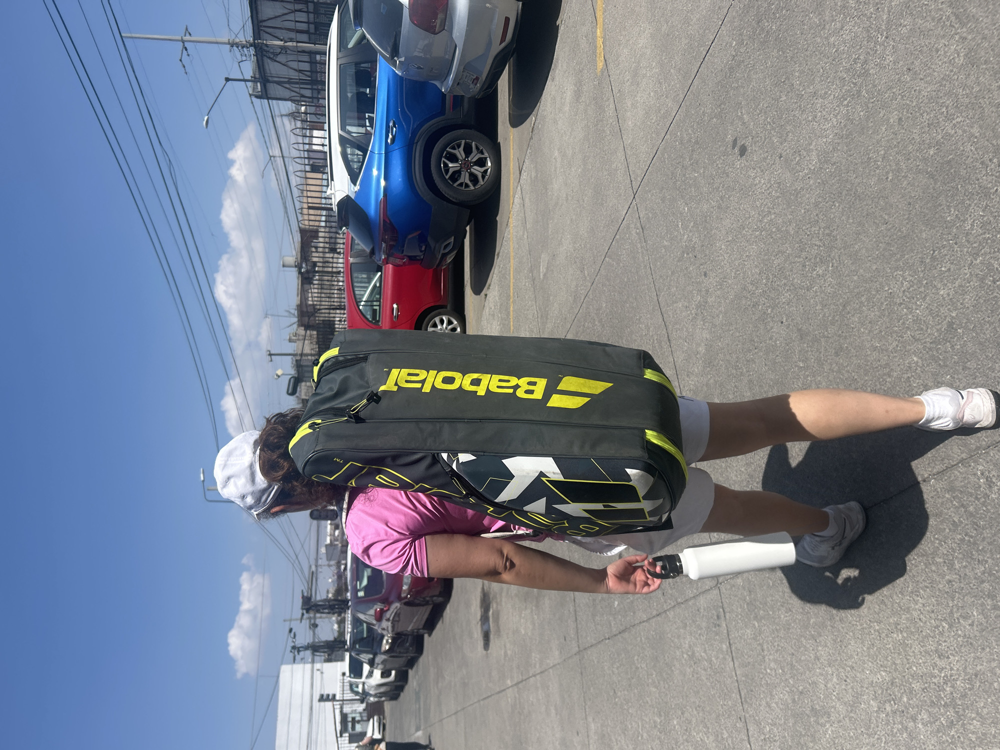 A person is walking outdoors on a paved surface near several parked cars. The person is wearing white shorts, a pink shirt, and white sneakers, and is carrying a large black and yellow Babolat tennis bag on their back and a white water bottle in their hand. The cars are a mix of colors, including red, blue, and silver. The sky is blue with some clouds. The scene is likely in Puebla, Mexico, based on the presence of overhead power lines and the type of parked cars.