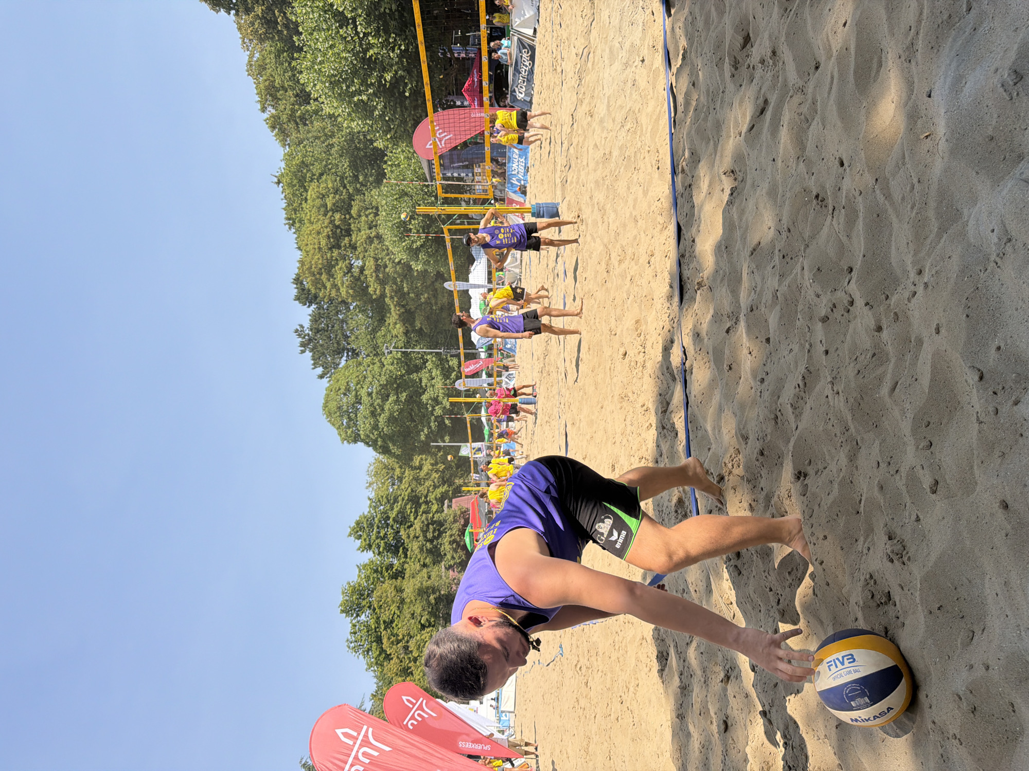 This is a medium shot, taken at eye level, of a beach volleyball tournament on a sunny day. In the foreground, a man wearing a purple tank top and black shorts is bending down to pick up a volleyball. The volleyball, a Mikasa Fivb Official Game Ball, is on the sand in front of him. In the background, several other people are playing volleyball, and a net stretches across the court. Red banners with white logos are visible on the left side of the frame, and trees form a backdrop behind the volleyball court. The scene appears to be outdoors, likely in a park or recreational area within Esch-sur-Alzette, Luxembourg, given the context.