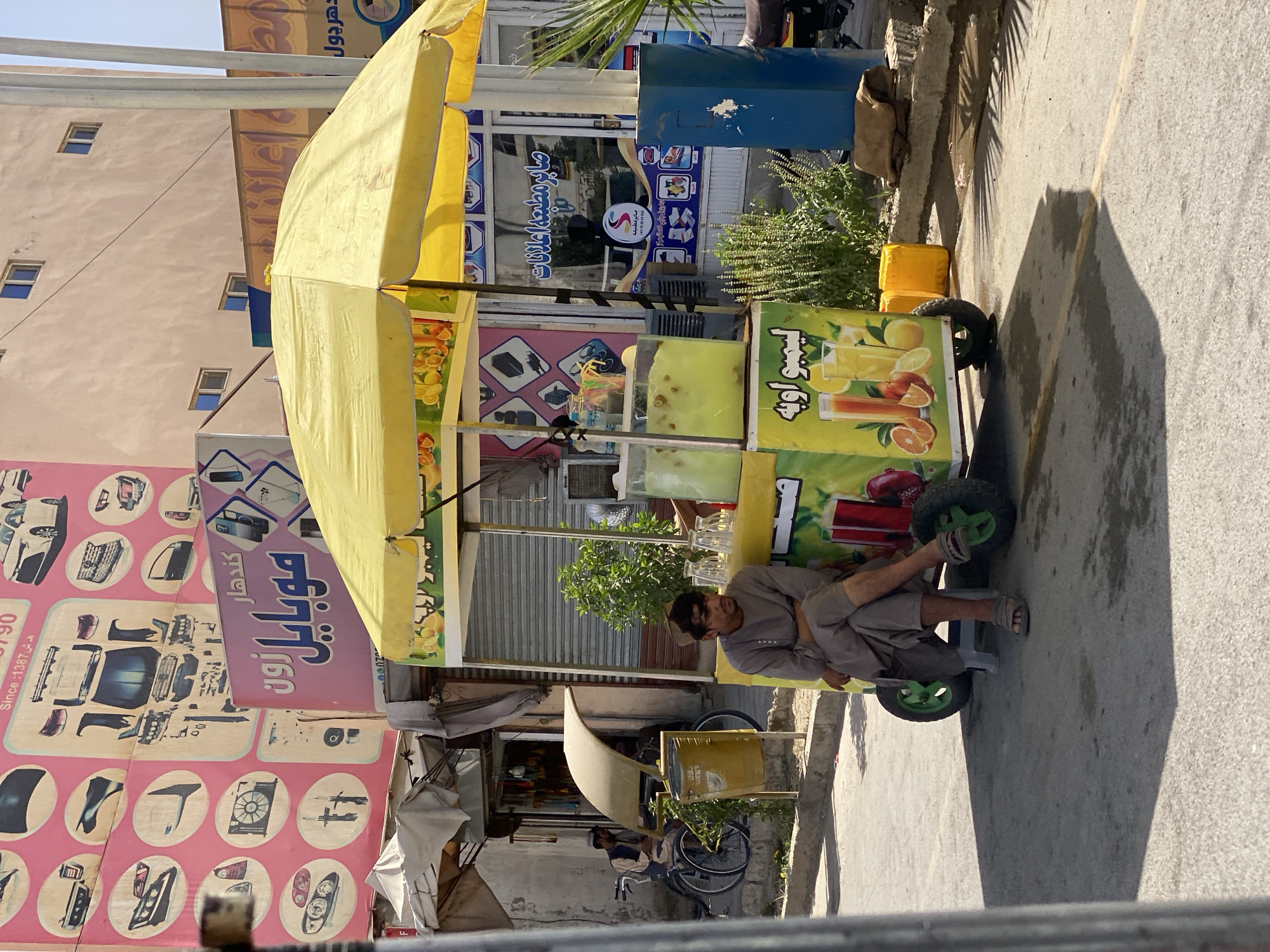 In Kandahar, Afghanistan, a man sits on a small plastic stool next to a brightly colored juice cart under a yellow umbrella. He is dressed in a grey shalwar kameez and wears sandals. The cart displays images of various fruits like oranges and lemons, with text in Arabic. To the left of the cart, a second man sits on a bicycle. Behind them, a building displays a large pink sign with images of car parts and text, and a purple sign with Arabic lettering that reads "Mobile Zone." The street is paved, and shadows from the umbrella and the surrounding structures create a dappled pattern on the ground. Several clear plastic cups and containers filled with yellow liquid are visible on the cart. The environment suggests a busy street scene in a city.