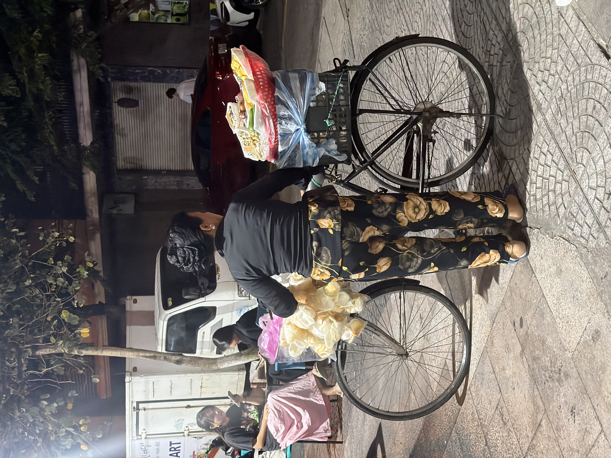 This nighttime scene, likely captured in Đà Nẵng, Vietnam, features a bustling street vending operation. The central figure is a woman, seen from behind, wearing a black long-sleeved top and dark pants with a distinctive yellow-orange floral pattern. She stands beside a bicycle fitted with a large cart, which functions as her mobile stall.
The bicycle cart is heavily laden with goods. In the front basket, a substantial pile of light-colored, thin, crispy snacks, possibly rice crackers or chips, is prominently displayed. Behind this, secured to the cart, are various bags of merchandise, including items in clear plastic packaging, a red mesh bag, and a blue plastic bag tied at the top.
Several other individuals are present. Partially visible behind the cart is another person, also in a black top, attending to items near the ground. Further to the left, a woman in a black shirt and a pink skirt sits on a low chair, engrossed in her phone. A young child or person is vaguely discernible near her.
The setting is an urban street or sidewalk, characterized by patterned paving stones. The dark background and reliance on artificial light indicate it is evening or night. Shadows are cast by the people and the bicycle. In the background, parked vehicles include a dark red car, a white truck, and a scooter. Green foliage from trees lines the left side of the image, and buildings with garage-style doors are visible further back.
Visible text on the side of the white truck includes "인천항" (Incheon Port in Korean) and "ART", along with partially visible Vietnamese text "cây món đồ!". The overall atmosphere suggests a typical evening street market or roadside vendor operation in a Southeast Asian city.