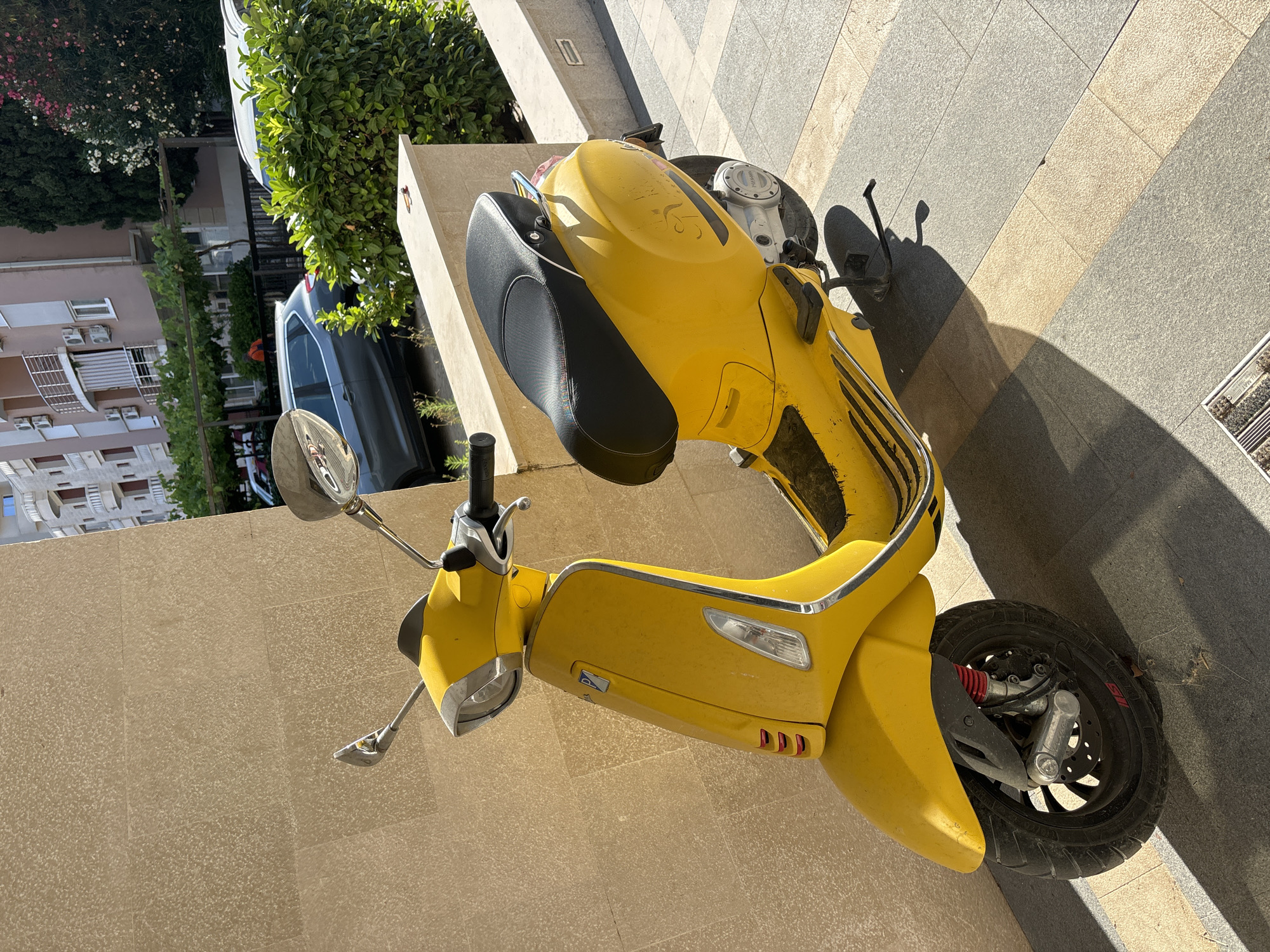 A yellow Vespa scooter is parked on a tiled walkway in Budva, Montenegro, casting a shadow from the bright sunlight. The scooter is angled towards the right of the frame, with its front wheel to the left. It has a black seat with a colored trim, chrome accents, and a visible kickstand. Behind the scooter, a light-colored building with balconies and air conditioning units is visible, along with lush green bushes and trees. Further in the background, a dark-colored car is parked. The ground is paved with rectangular tiles of varying shades of gray and beige.