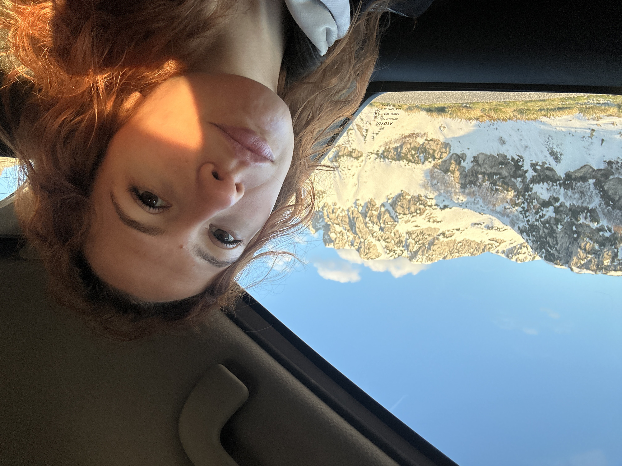 The image depicts a woman with reddish-brown hair, partially visible in the upper left, looking toward the camera from inside a car. The scene outside the car window includes a mountainous landscape with snow-covered peaks and a clear blue sky. Based on the mountainscape, the location is indicated as Pašina voda, Montenegro. The vehicle's interior, including the dashboard and window frame, is also visible in the foreground. There is a white cloud behind the mountains.