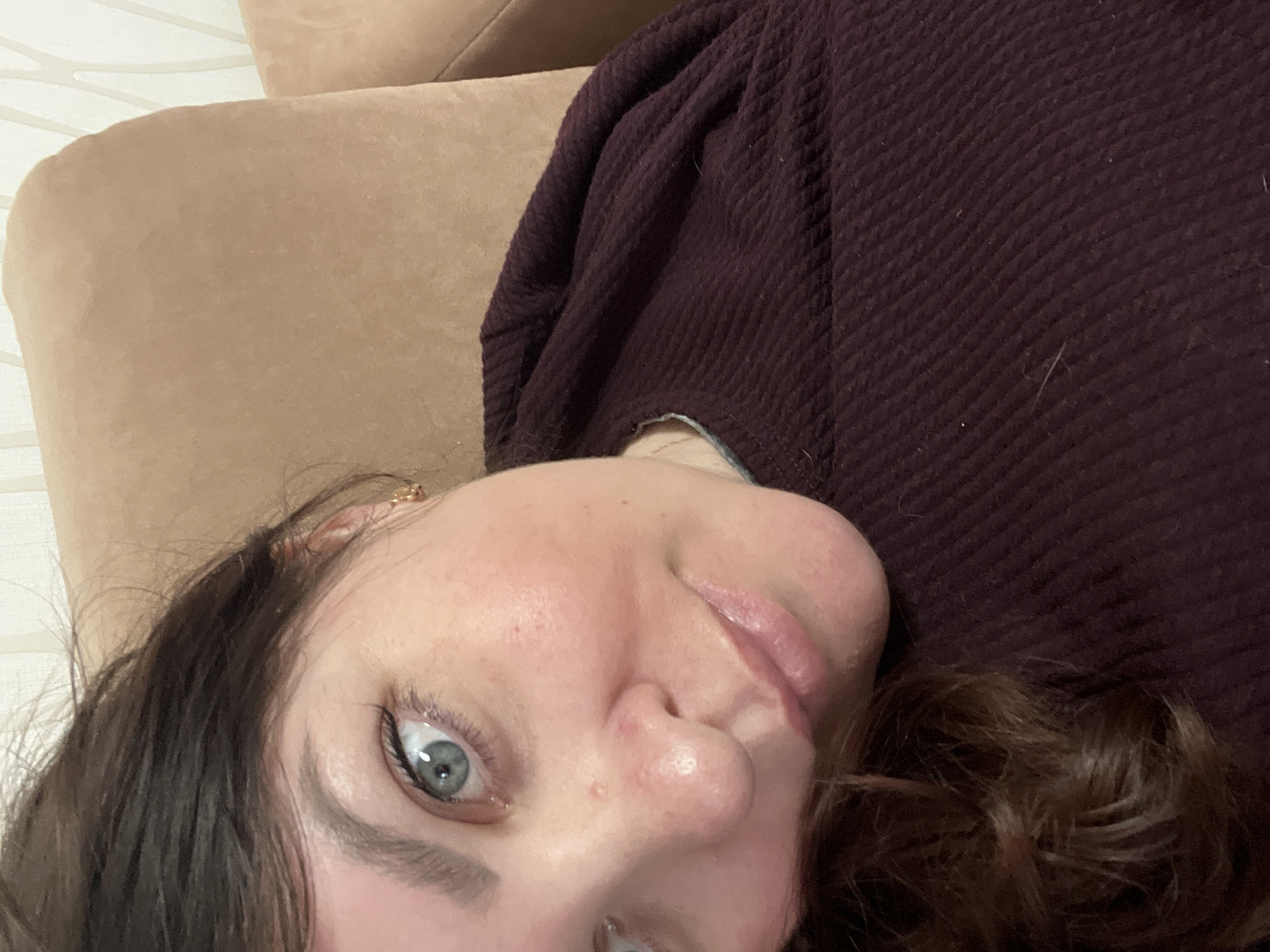 The image is a close-up, high-angle portrait of a young woman positioned horizontally, looking upwards at the camera. Her head is tilted to the side, causing the image to appear rotated. She has fair skin, dark brown wavy hair, and striking light blue or green eyes, which are framed by dark, well-defined eyebrows. She wears subtle makeup, including black eyeliner and a glossy pink lip. A small, delicate golden earring is visible on her left ear.
The woman is wearing a dark maroon or burgundy long-sleeved garment, which features a distinct textured, possibly waffle-knit, pattern. Light specks, resembling lint or dust, are visible on the fabric. She is lying on a light brown or beige upholstered surface, likely a sofa or armchair, which occupies most of the background. Behind this furniture, a light-colored wall with a subtle, faintly patterned texture is visible.
The setting appears to be indoors, suggesting a comfortable, domestic environment. Her expression is calm and direct, with a gentle gaze towards the viewer, conveying a relaxed and intimate mood. The lighting is soft and even, typical of indoor conditions. No specific time of day or weather conditions are discernible, nor is any visible text present in the image. The location context for the image is Perm, Russia.