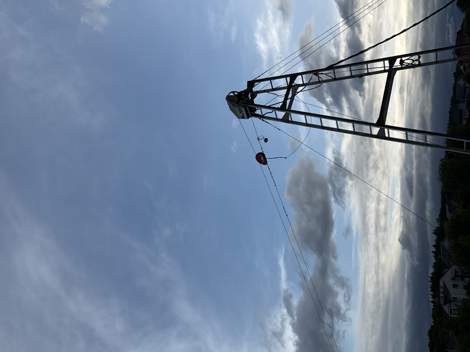 This media file presents a dramatic sky scene featuring a large industrial structure. The image, when correctly oriented, shows a prominent, lattice-like metal boom or crane segment positioned on the right side, extending diagonally upwards towards the top-left. Cables and wires are strung from this structure, and a small, red, oval-shaped object is suspended from one of the wires. The metal structure prominently displays the vertical inscription "WOCIE".
The sky exhibits striking weather conditions, likely indicating late afternoon or early evening. Patches of clear blue sky are visible, predominantly on the left, while the right and upper portions of the frame are filled with an impressive array of clouds. Some clouds are dark and heavy, suggesting potential storm activity, while others are brightly illuminated by sunlight, creating a high-contrast and moody atmosphere.
Along the distant horizon line, visible beneath the clouds on the bottom right, are silhouettes of trees and residential buildings, indicating a semi-urban or suburban setting. No human activity or specific event is taking place; the scene is static, focusing on the interplay between the industrial structure and the dynamic sky.