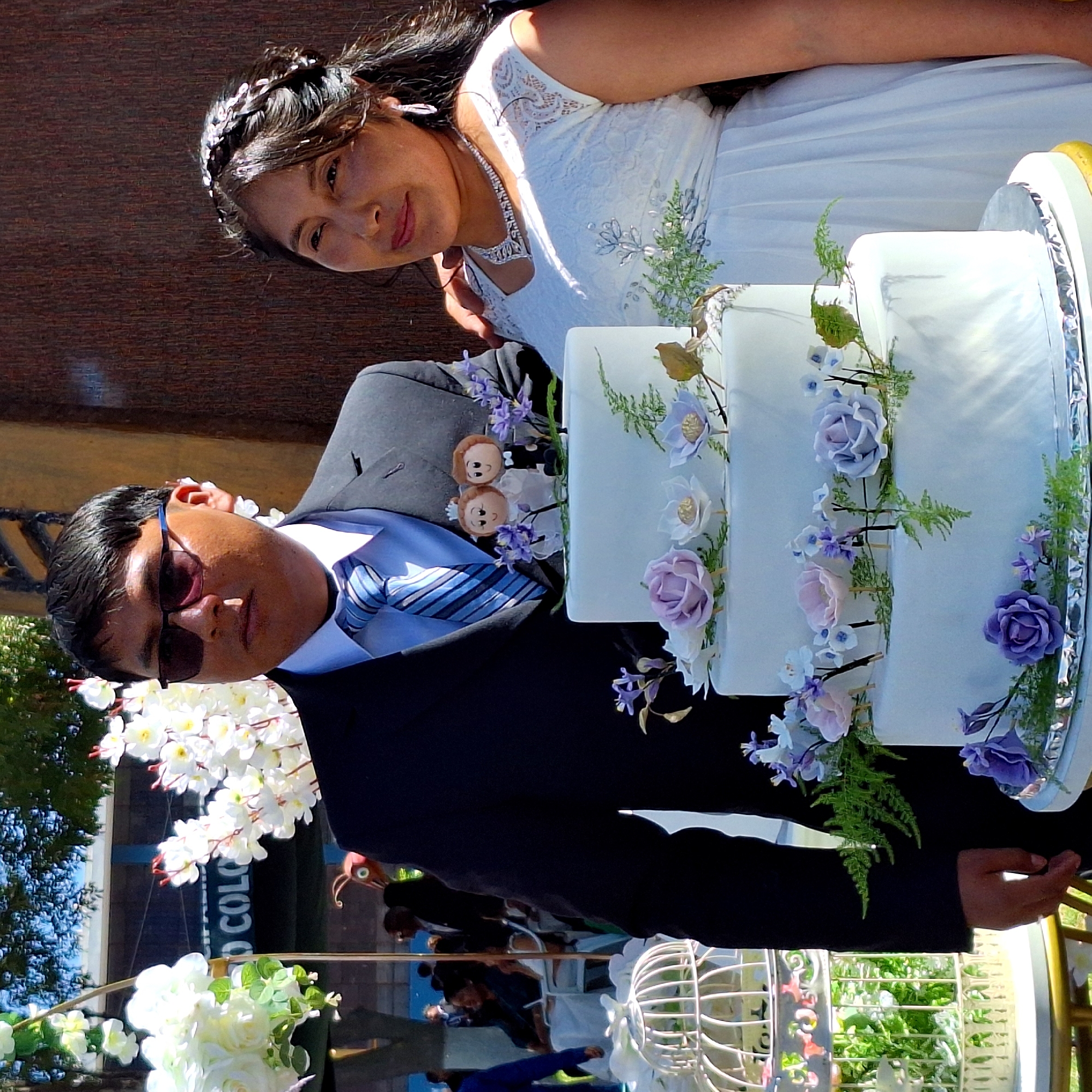 The image shows a wedding celebration with a bride and groom posing next to a three-tiered wedding cake decorated with flowers. The groom is wearing a dark suit with sunglasses and a blue tie, while the bride is in a white dress with a tiara and necklace. The location appears to be outdoors, possibly in a garden or patio, with a glimpse of building signage that indicates it may be in Cerro Colorado, Peru. There are other people in the background, along with decorative flowers and birdcage decorations.