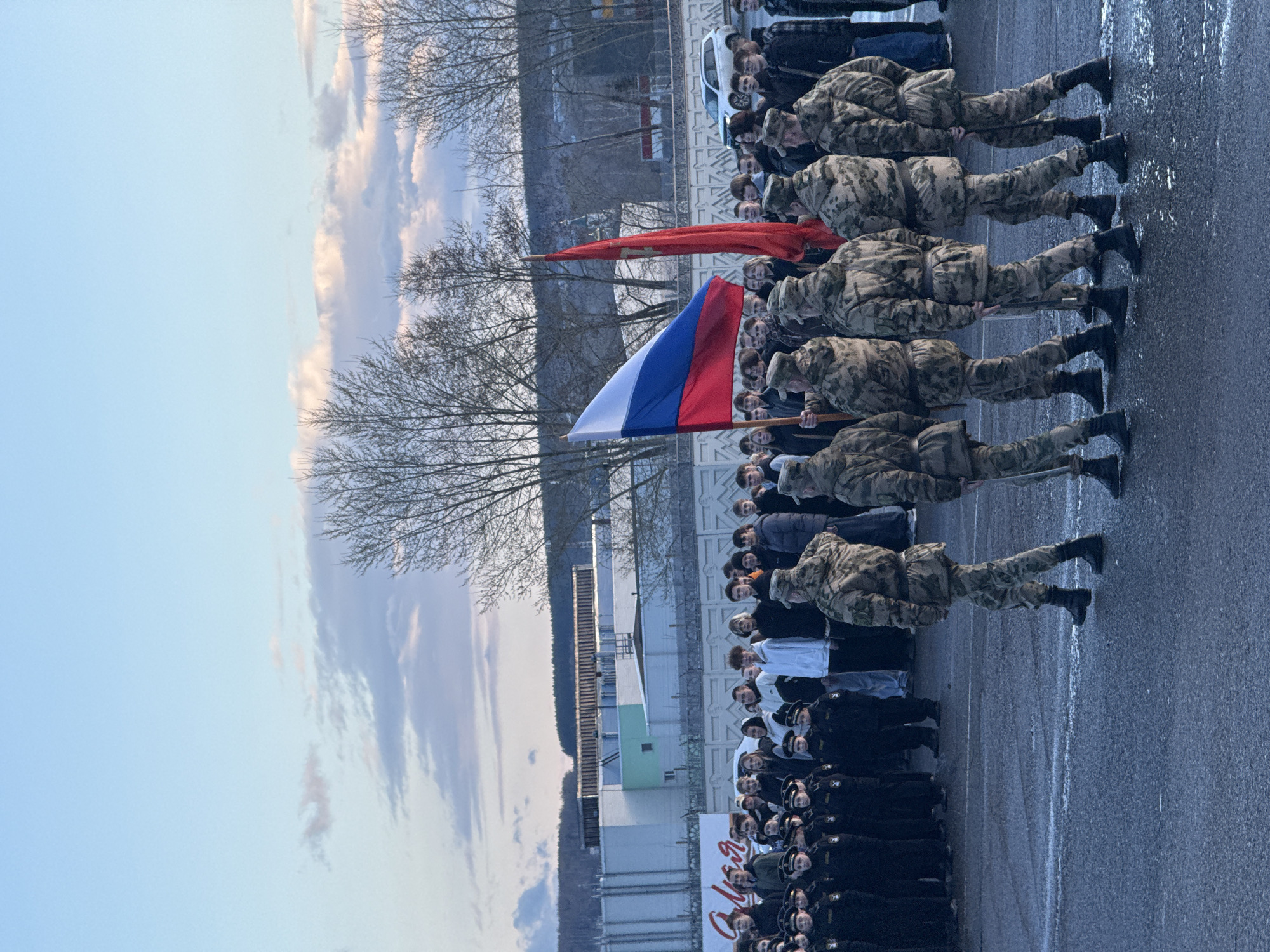 An outdoor ceremony or gathering is captured from a slightly elevated perspective on what appears to be a paved area. In the foreground and middle ground, a line of at least five individuals dressed in camouflage military-style uniforms stands at attention, each holding a pole. Two prominent flags are displayed: the Russian national flag (white, blue, and red horizontal stripes) and a solid red flag, possibly a Soviet or military banner.
Behind the uniformed participants, a large crowd of spectators or participants is gathered. This group includes individuals in dark uniforms, likely students or cadets, and others in civilian clothing. More people are visible in the background, near a white car.
The scene features bare trees on the left and in the background, indicative of a colder season such as late autumn, winter, or early spring. A light-colored building with a green facade is visible to the left. The sky is overcast with light clouds, suggesting a daytime event. The ground appears damp or wet, possibly from rain or melting snow. In the far background, the faint outline of hills or mountains is visible.
A white banner or sign in the mid-left background displays partially visible Russian text, starting with "Я" followed by cursive script that resembles "Мая" (May), suggesting the event may be related to May Day or Victory Day. The overall atmosphere conveys a formal, possibly patriotic or commemorative occasion taking place in Votkinsk, Russia.