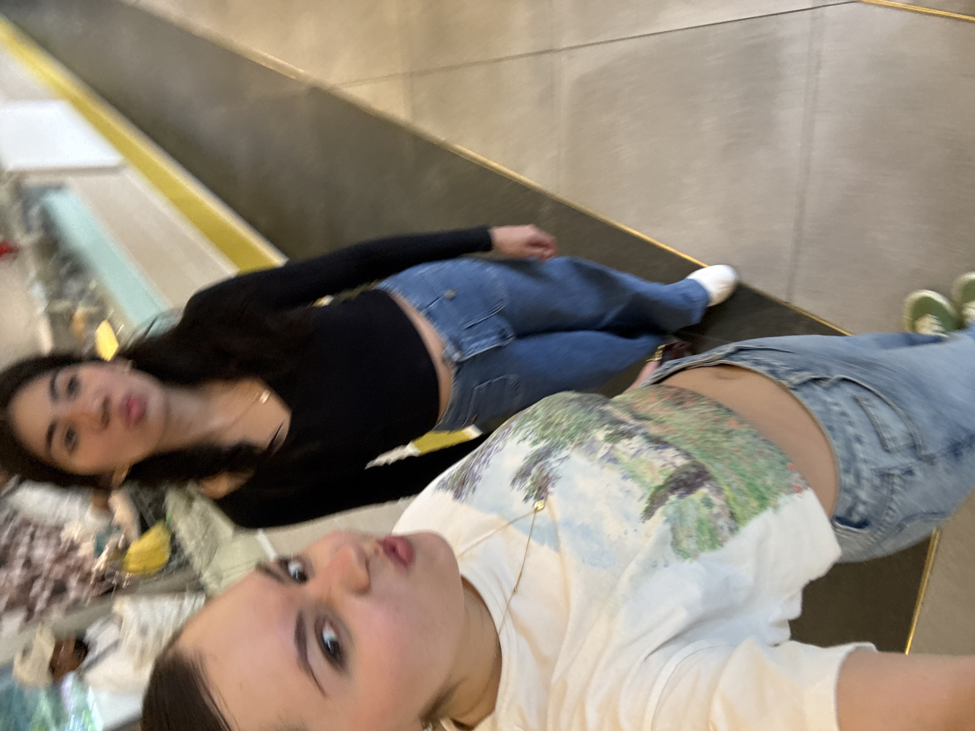Two young women are lying down, posing for a selfie. The woman closest to the camera is wearing a white t-shirt with a nature-themed design and light-wash jeans. She is also wearing a gold necklace. The other woman is wearing a black long-sleeved shirt and blue jeans. She has dark hair. Both women are wearing casual footwear. They appear to be lying on a ledge or platform with a black top and a gold trim in an indoor space. The location may be the Museo Soumaya in Mexico City, Mexico, given the architectural features of the surrounding structures.
