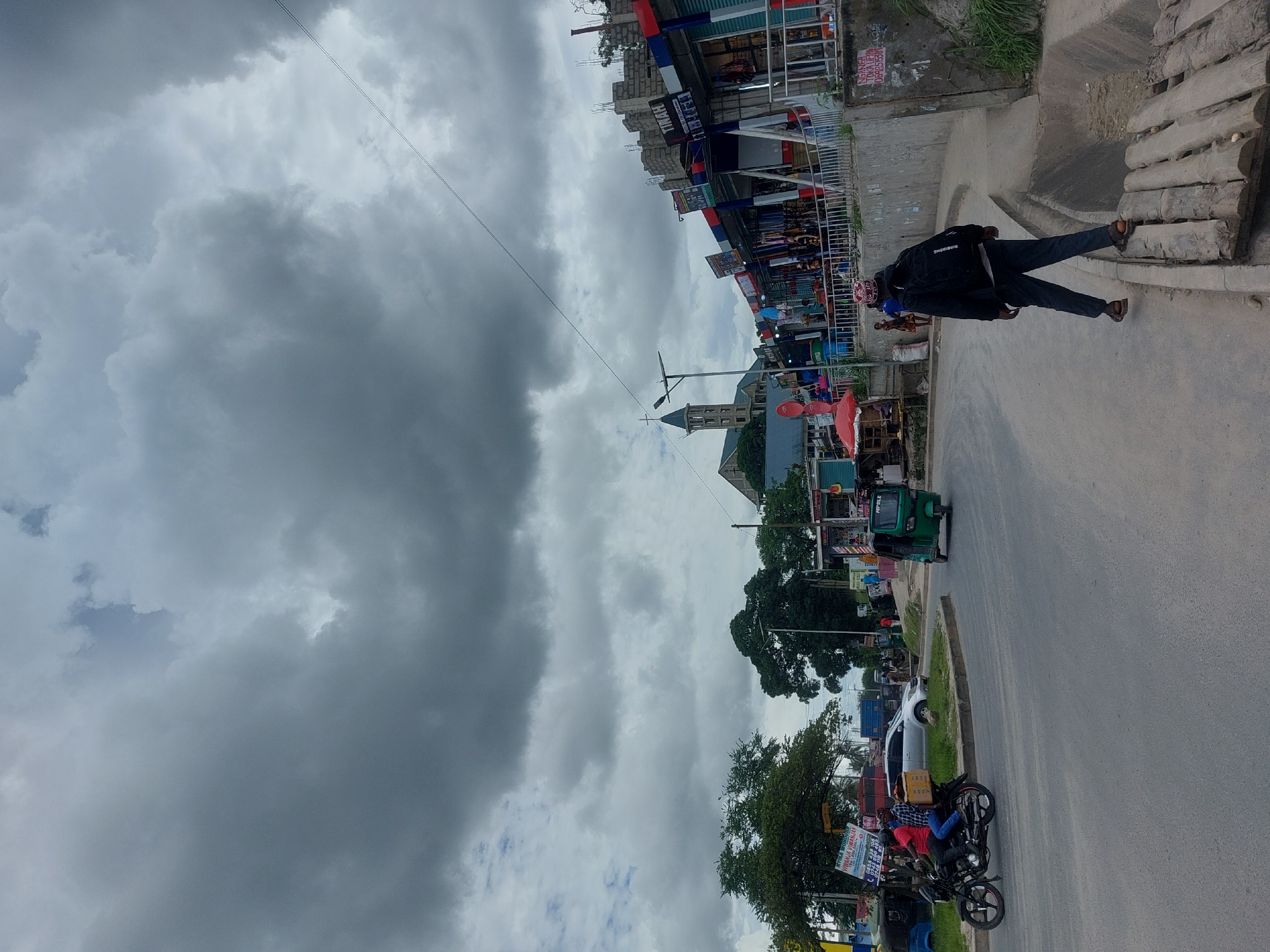 Here's a detailed description of the image:
**General Overview:** The image depicts a street scene under a heavily clouded sky. It appears to be taken in a bustling area.
**Location:** The presence of Swahili signage and architecture suggests the location is Dar es Salaam, Tanzania. The mix of buildings, including what appears to be a church with a prominent spire, is typical of the city.
**People:** Several people are visible. One man is standing on the edge of the sidewalk, wearing a dark jacket and trousers. He has a patterned helmet and sandals. Further down the street, two people are seated on a parked motorcycle with signage attached.
**Objects:** Several objects are visible:
* A three-wheeled vehicle (Tuk-tuk) is present.
* Several buildings are visible, appearing to be a mix of shops, residences and religious structure.
* Various signs and advertisements are displayed along the street.
* Power lines are crossing the sky.
**Actions:** The main action appears to be everyday street activity.
**Interactions:** The main interaction appears to be everyday street activity, including pedestrian and vehicle traffic.
**Environment:** The sky is dominated by heavy, dark clouds, suggesting possible rain. The street surface is paved, with sidewalks on both sides. There are also trees and greenery along the roadside. The image suggests a dense urban environment.