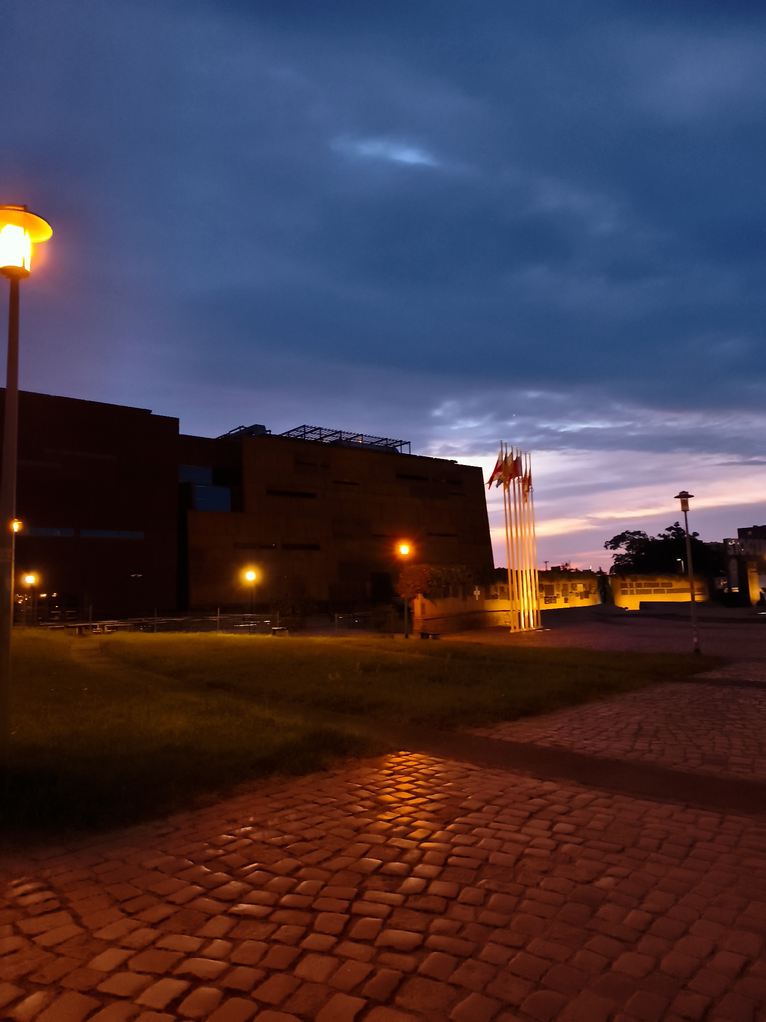 The image captures a twilight scene outside a modern, dark-colored building in the city of Gdańsk, Poland. A cobblestone path, illuminated by warm streetlights, leads towards the foreground. To the left, a lamppost with an orange glow stands near a grassy area. The building features large, dark windows and an industrial-looking rooftop structure. In the middle ground, a cluster of tall flagpoles, lit from below, stands prominently, adorned with several flags. The sky above is a deep blue, transitioning to lighter shades with wisps of clouds reflecting the last light of the day. The overall atmosphere is one of dusk and calm.