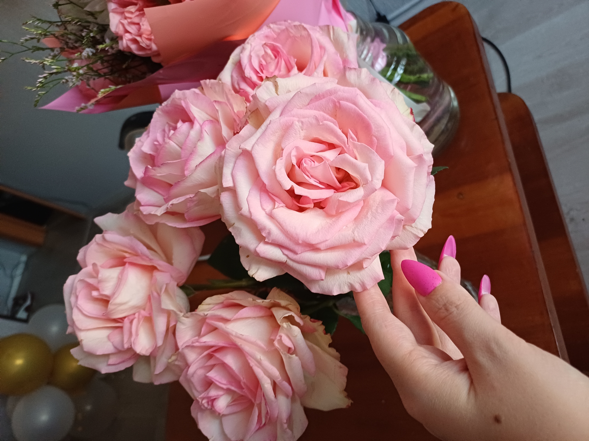 A close-up shot captures a bouquet of light pink roses with some white and darker pink edges, arranged in a glass vase on a wooden surface. A hand with bright pink, stiletto-shaped nails gently touches one of the rose petals, drawing attention to the delicate texture of the flowers. In the background, a hint of other floral arrangements can be seen, along with some balloons in gold and gray. The lighting suggests it might be indoors, possibly in the evening or a dimly lit room, with the warm tone of the wood and the soft lighting creating a serene and romantic atmosphere. The focus is on the beauty of the roses and the touch of human interaction, conveying a sense of appreciation for nature's artistry. The overall mood is gentle and intimate. There is no visible text in the image.
