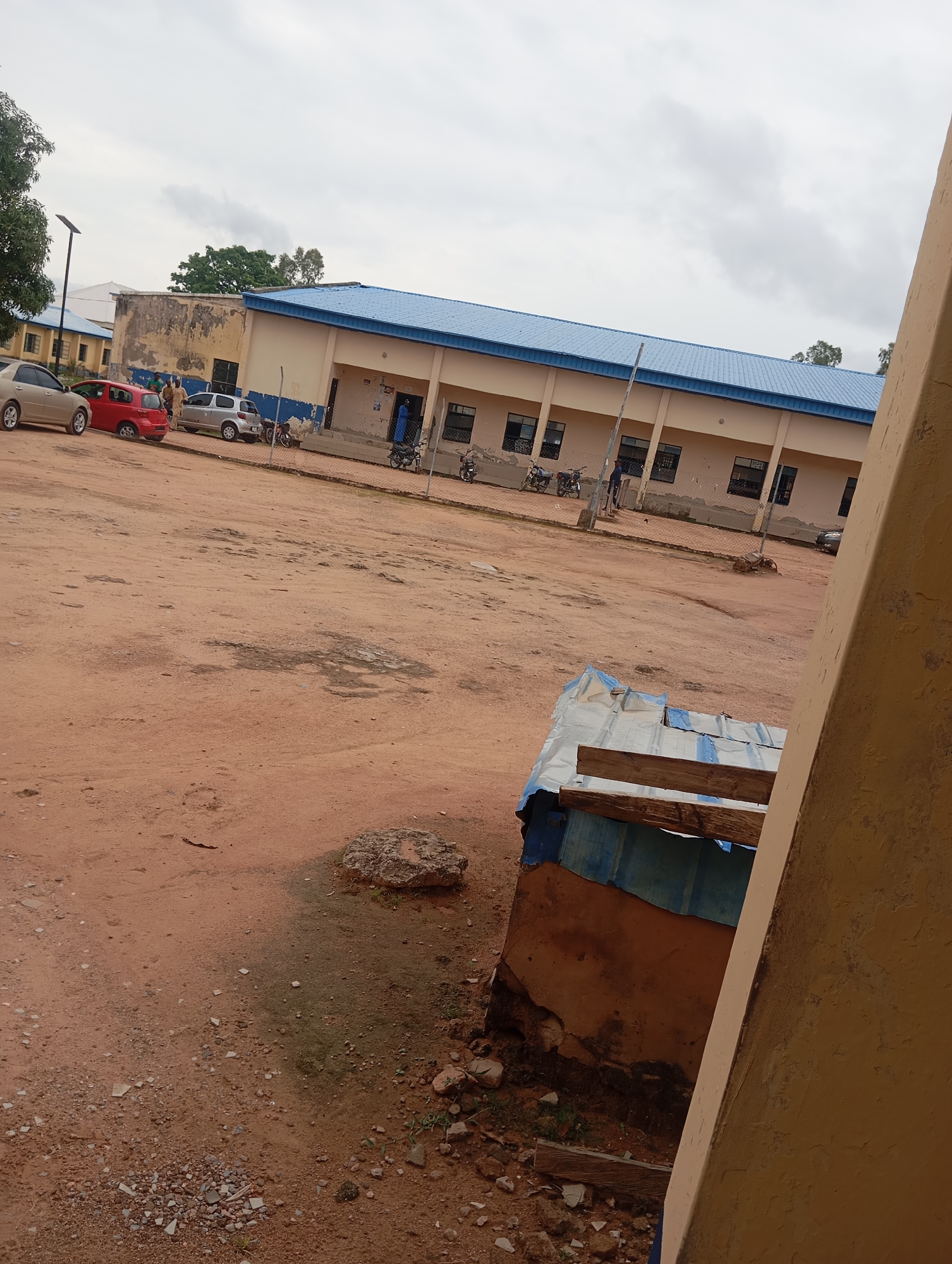 This image captures an exterior scene in Jalingo, Nigeria, likely a school or public building complex. The foreground is a dusty, reddish-brown dirt ground with scattered pebbles and debris. To the left, parked cars are visible: a gold sedan, a red hatchback, and a silver SUV. Several motorcycles are parked near the building.
The main subject is a long, single-story building with a blue corrugated metal roof and cream-colored walls. The building features a row of dark-framed windows and several doorways. A chain-link fence runs along the front of the building, separating it from the open area. In the middle ground, a makeshift shelter with a corrugated metal roof and blue plastic sheeting is partially visible, appearing weathered.
The sky is overcast with grayish clouds. A single solar-powered street lamp is visible in the distance on the left. A few trees are sparsely scattered in the background. The overall impression is of a utilitarian, somewhat aged environment.