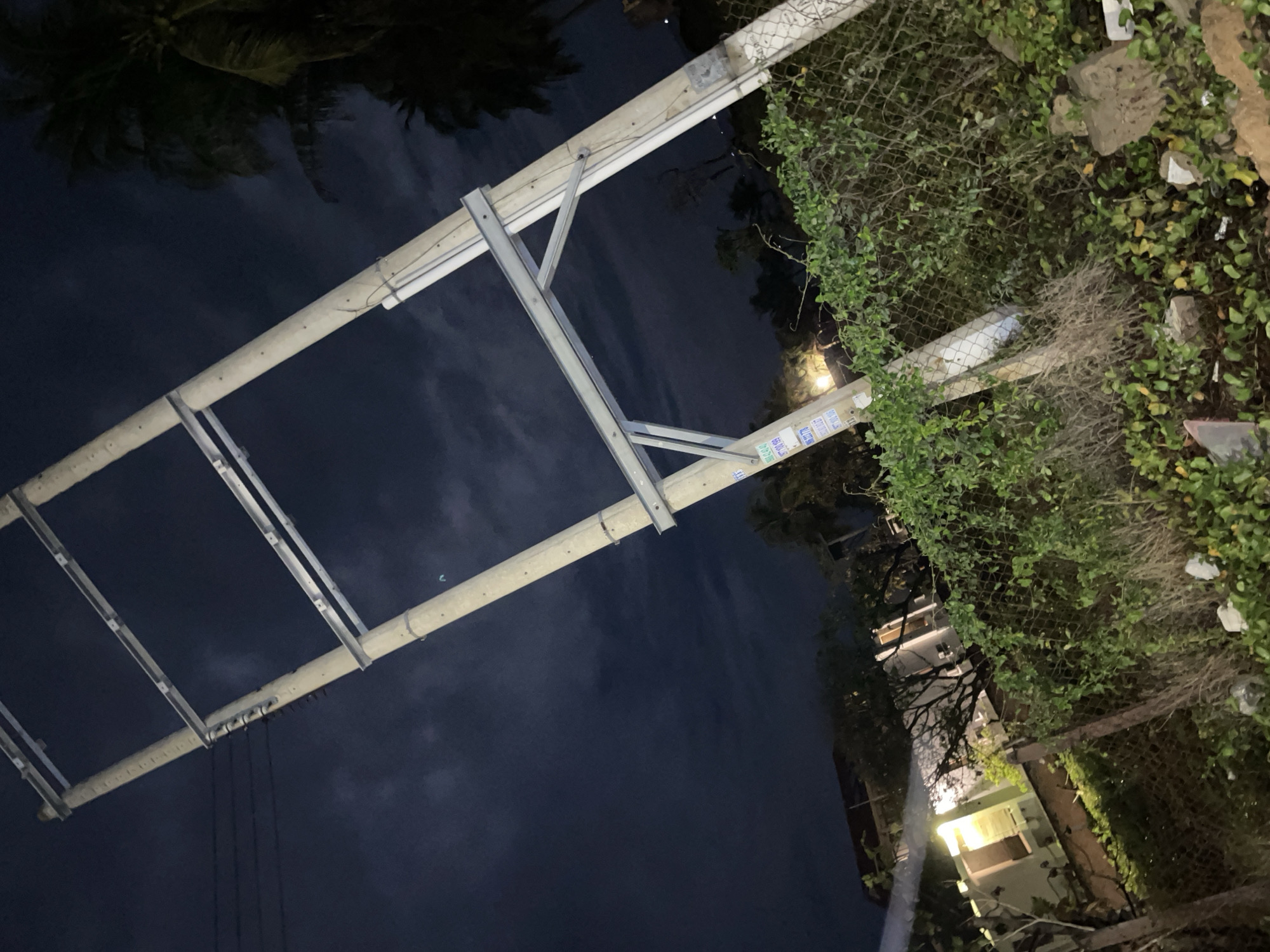 A utility pole with metal support beams stretches diagonally across the frame against a dark sky, suggestive of Phu Thuy Ward, Vietnam. The pole is situated next to a chain-link fence heavily overgrown with vines and foliage. Beyond the fence, a building with illuminated windows can be seen. Electrical wires are visible extending from the pole. Palm tree fronds are visible in the upper left corner. Debris is scattered around the base of the fence.