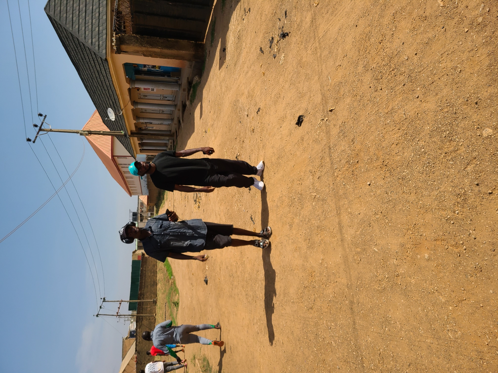 The image depicts an unpaved dirt road in what appears to be a residential or commercial area of Jos, Nigeria, based on the architectural styles of the buildings and the presence of utility poles. Two adult males are walking along the road. One is wearing a black shirt and pants with white sneakers and a blue cap. The other is wearing a denim-like shirt, dark shorts, sandals, and what appears to be headphones. There are other people further down the road, some of whom appear to be exercising or stretching. The environment is dry and dusty. Buildings with varying architectural styles are visible along the side of the road.