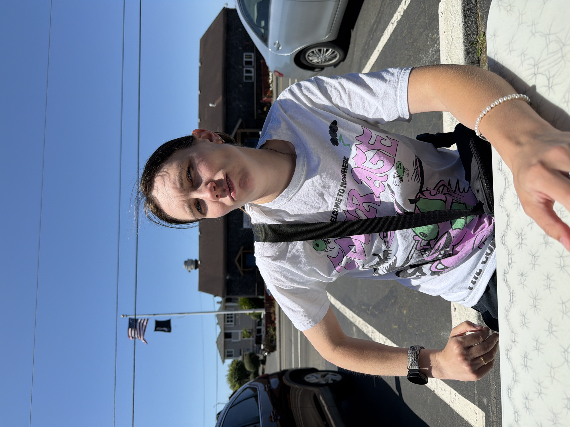 The image captures a person sitting at an outdoor table, likely at a restaurant or cafe, on a sunny day in Cannon Beach, Oregon, United States. The background reveals a street scene with parked cars, buildings, and a flagpole flying the American flag and another flag below it. The person, who appears to be a young woman, is wearing a white t-shirt with a graphic print and a black crossbody bag. She has a watch on her left wrist and a bracelet on her right wrist. Her expression is directed towards the camera. The foreground features the edge of a white patterned table. The overall environment suggests a casual outdoor setting with a clear blue sky overhead.